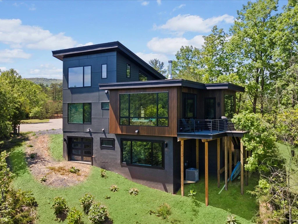  Mountain house architect project featuring gray siding, wood accents, large windows, and a raised balcony on a wooded hillside.