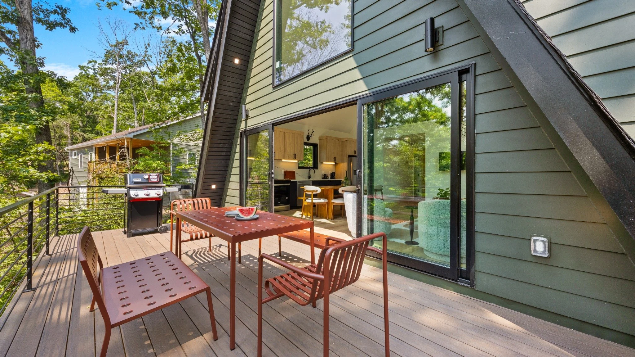 Residential architect project featuring green siding, black-framed glass doors, and a wood deck with red dining furniture and a grill.