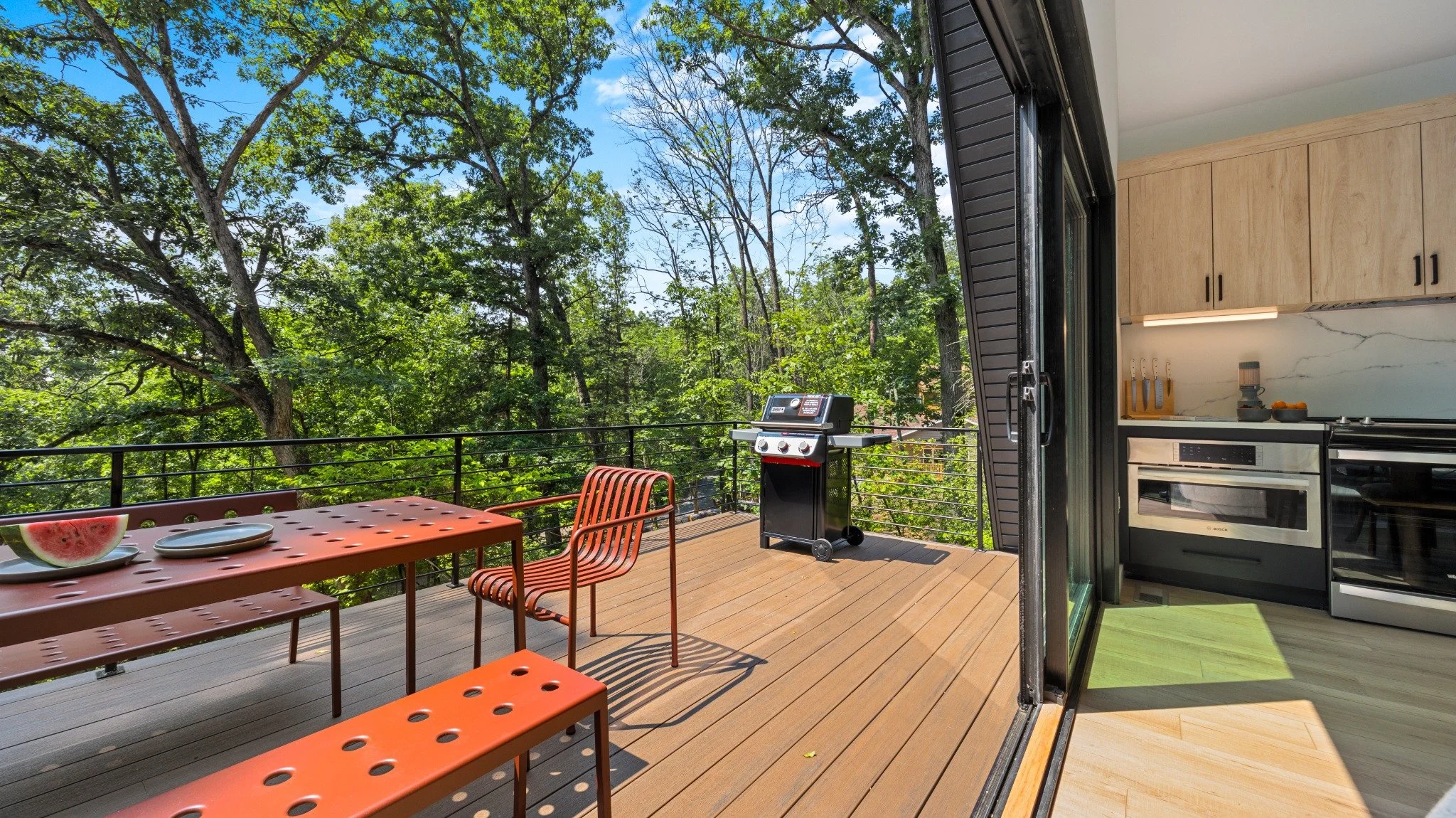 Custom home designer project featuring sliding glass doors connecting a wood-cabinet kitchen to a deck with red dining furniture and a grill.