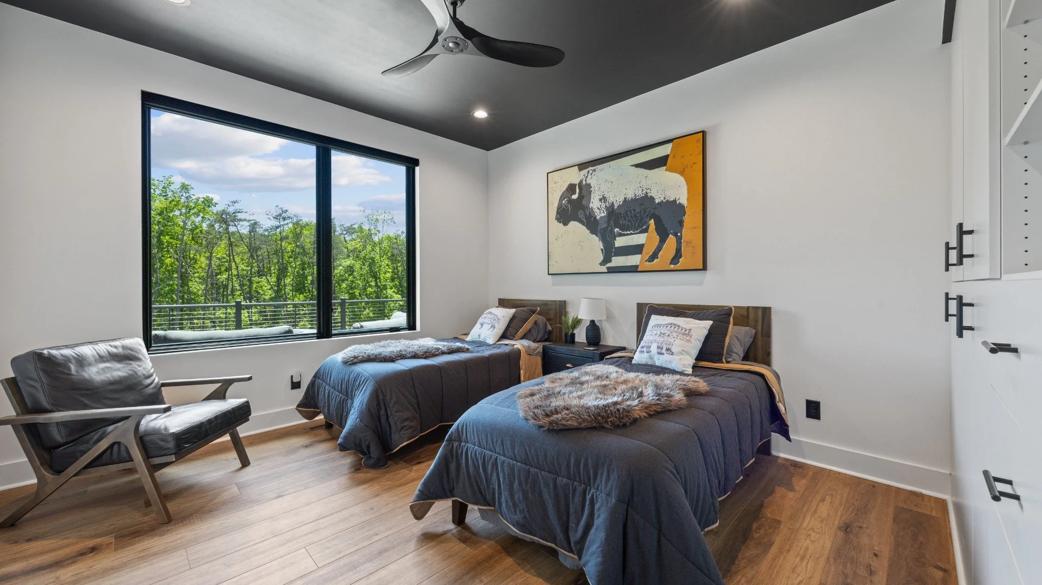 Custom home designer project featuring twin beds, dark bedding, a black ceiling, a leather lounge chair, and a large window overlooking trees.