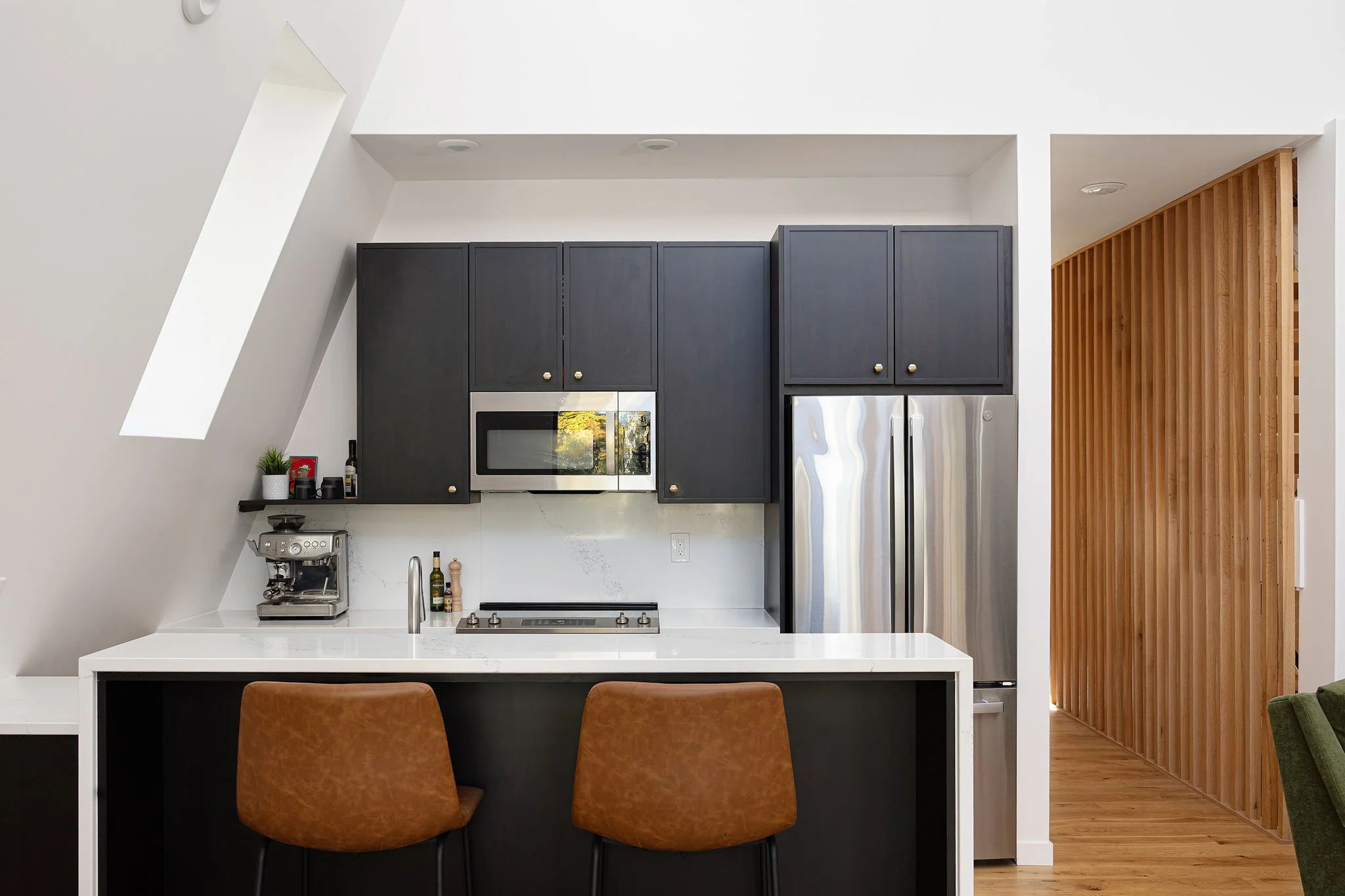 Modern kitchen with black cabinetry, stainless steel refrigerator and microwave, white marble countertop, and wooden slat accent wall.