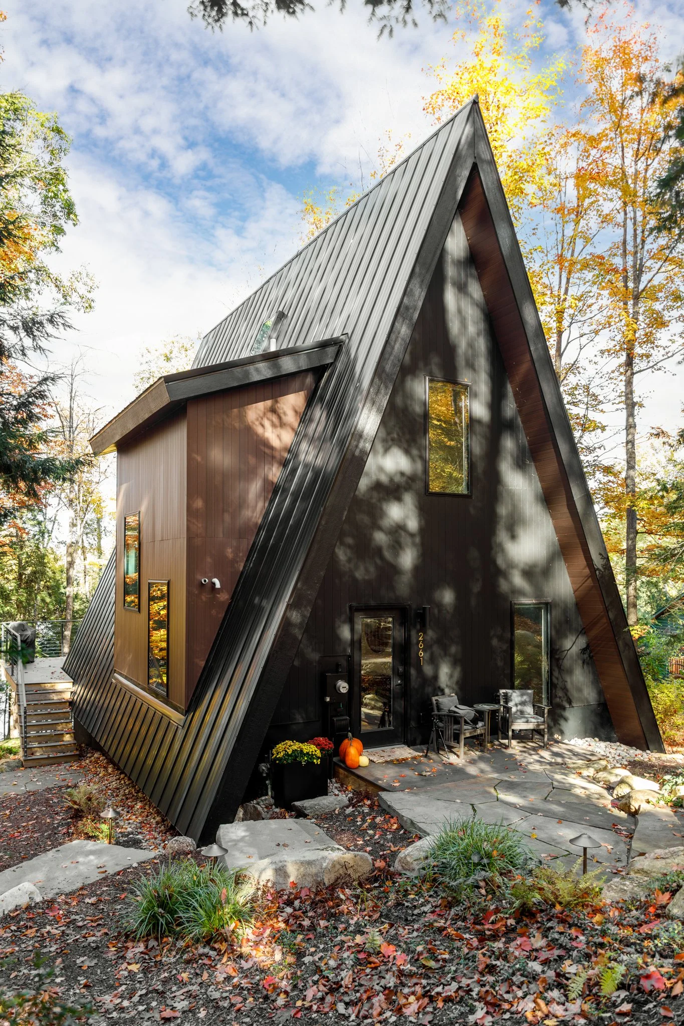 Modern A-frame house with dark exterior, large windows, surrounded by autumn trees and fallen leaves.