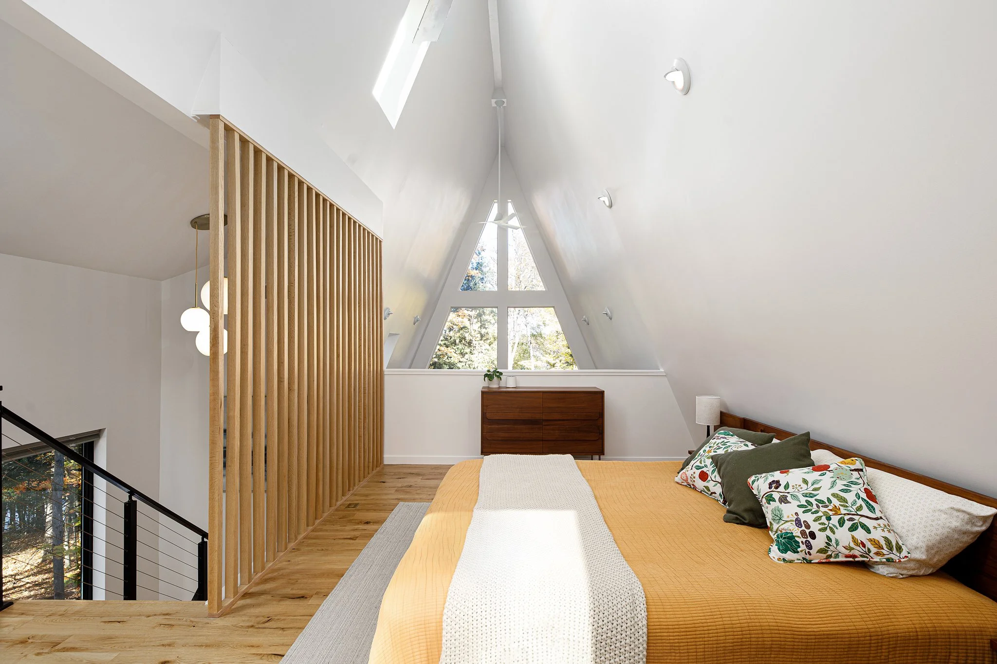A bedroom with a sloped ceiling, a large window, a wooden dresser, and a bed with pillows and a beige blanket. The room has white walls, wooden floors, and modern lighting.