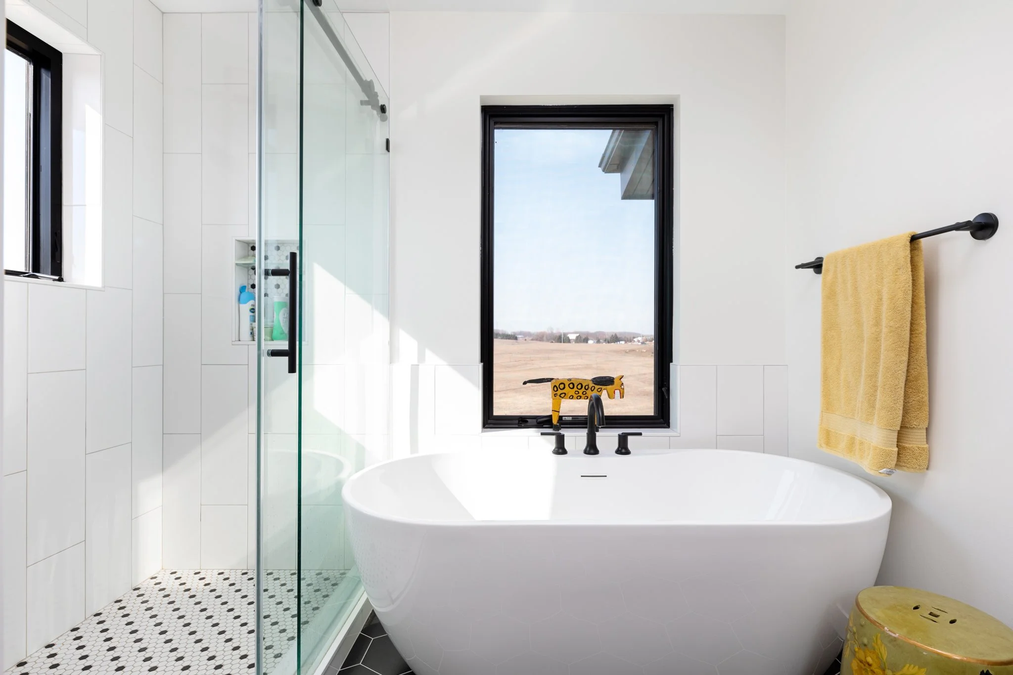 Modern white bathroom with a freestanding bathtub, black faucet, yellow towel on a black towel bar, window showing an outdoor field, and walk-in shower with glass door.