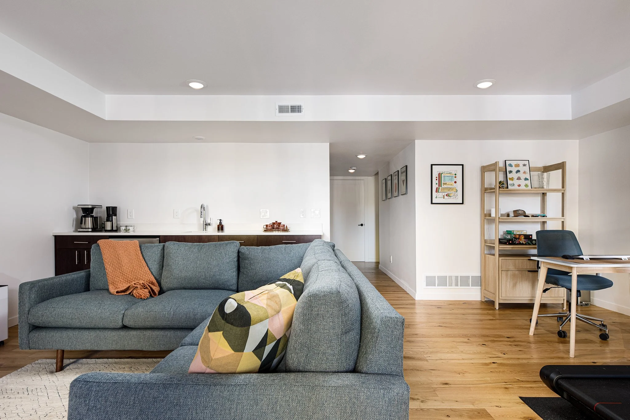 Living room with gray couch, wood flooring, small desk with blue chair, white walls with framed artwork, and a small kitchen area in the background.