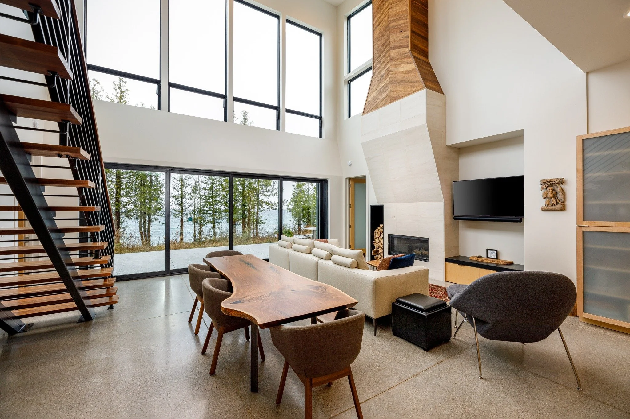 Lakefront home design with a wood dining table, white sofa, open wood-and-metal staircase, tall fireplace, and water views.
