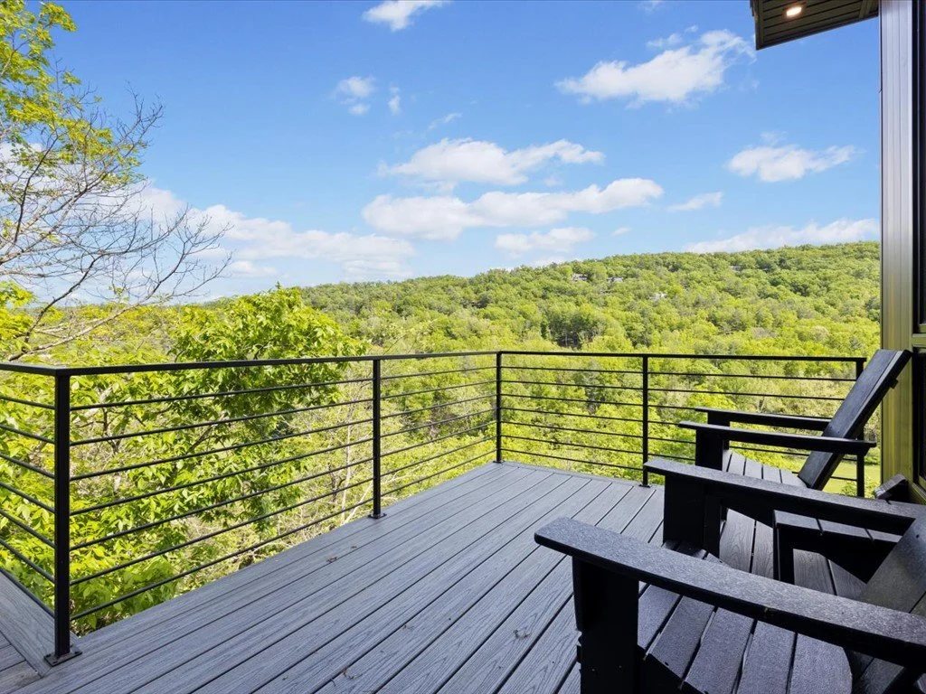  Mountain home architect project featuring black Adirondack chairs, a gray deck, and metal railings overlooking wooded hills.
