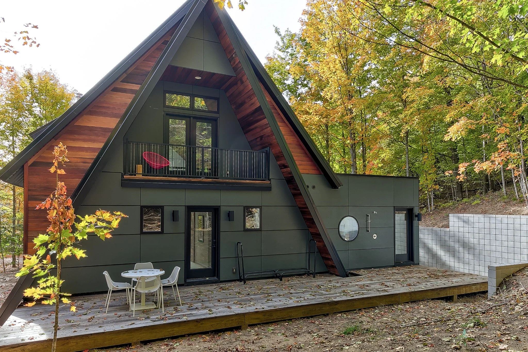  Contemporary lake house design with a steep roof, wood accents, red doors, and a stepped deck among autumn trees.
