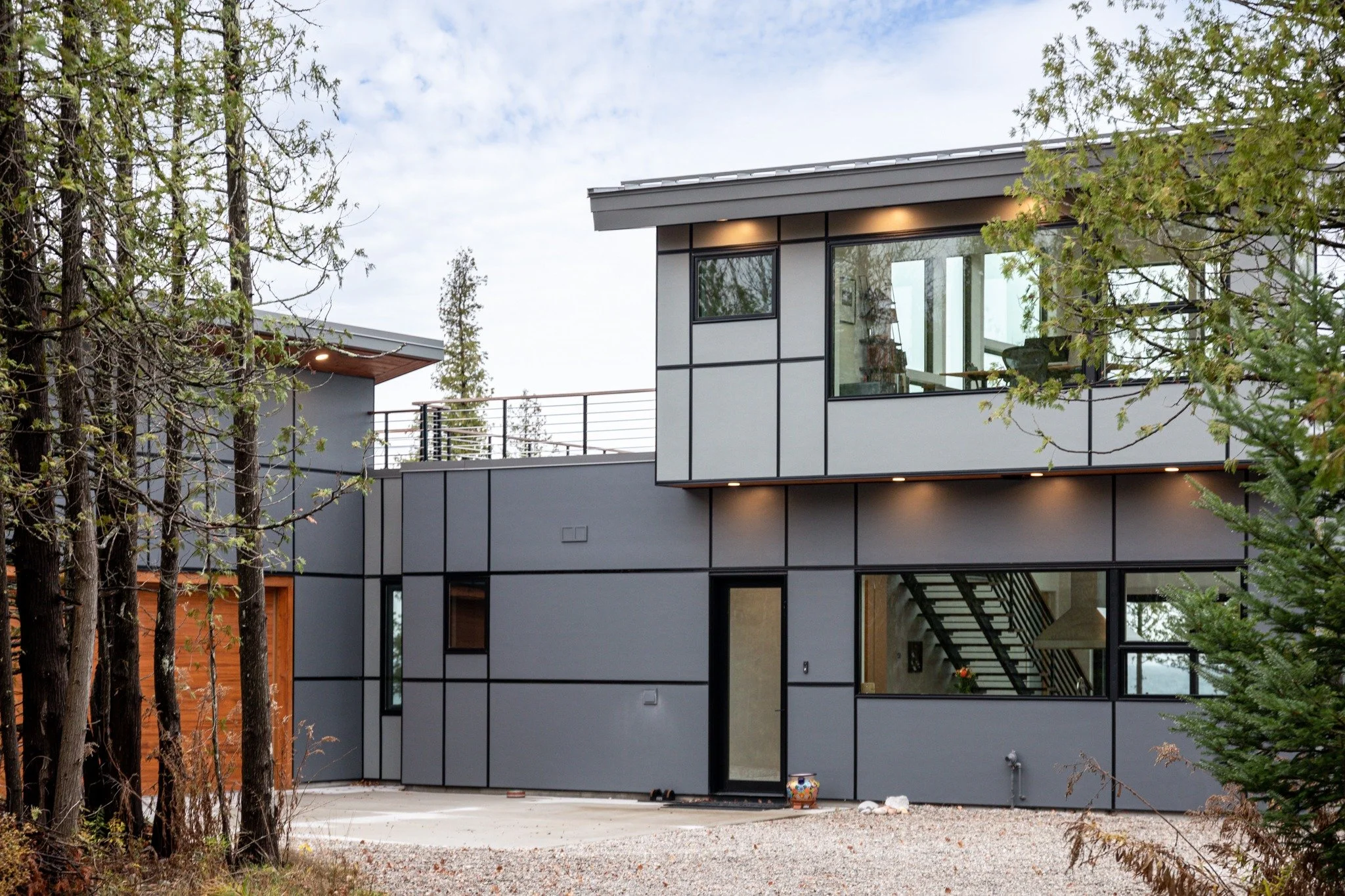 Lake house exterior design with gray panel siding, large black-framed windows, an upper terrace, and evergreen trees.
