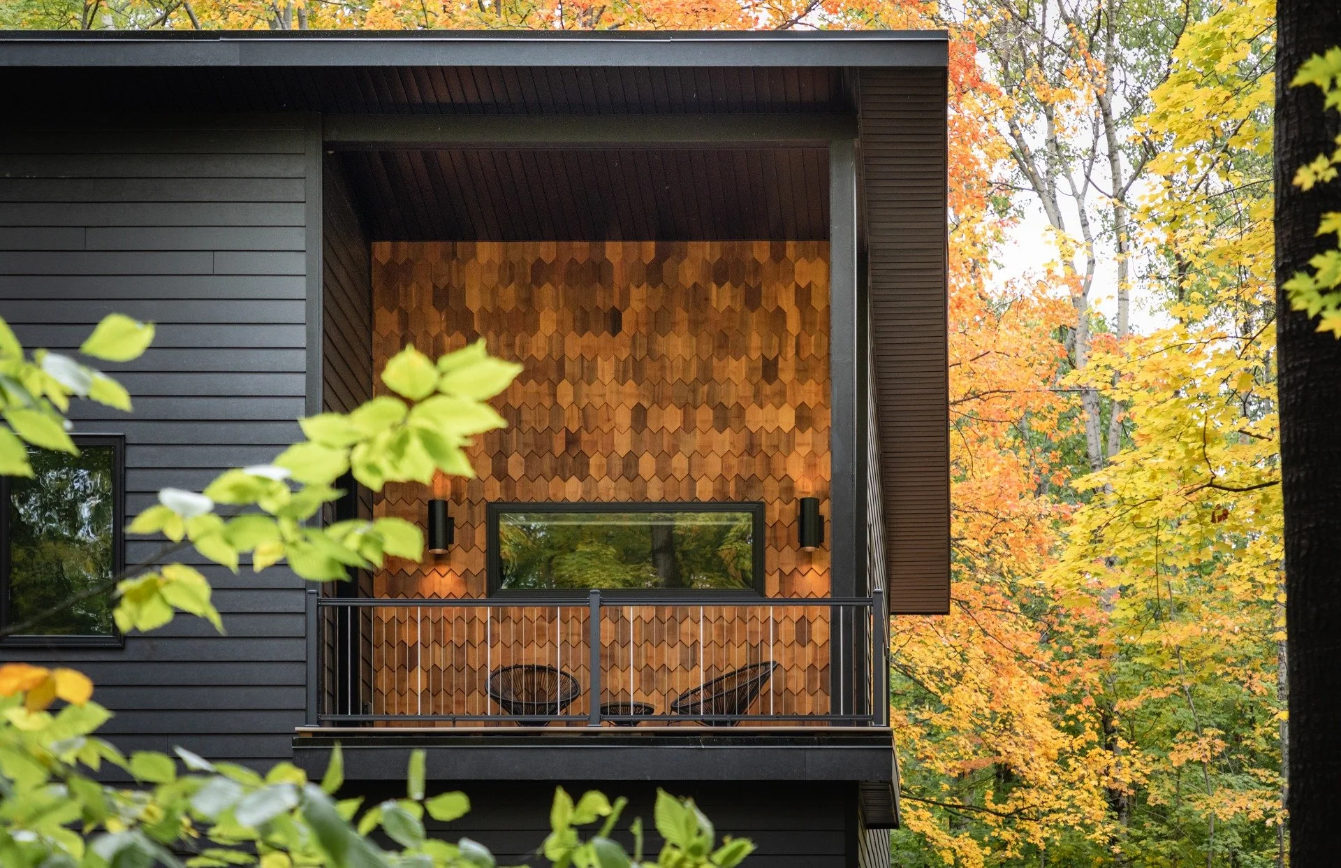 Custom mountain home architect project featuring a covered balcony, wood-shingle accent wall, dark siding, and black railings framed by autumn leaves.