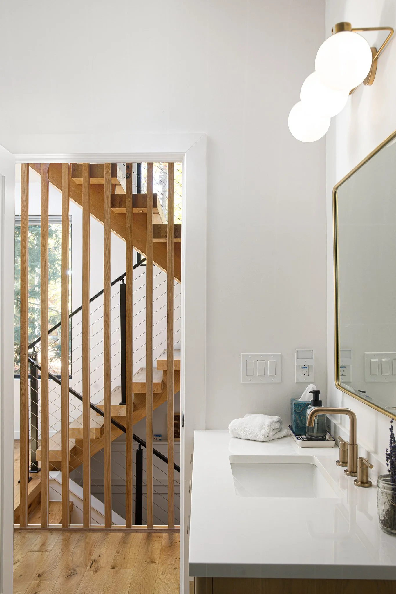 A modern bathroom corner with a white countertop, a brass faucet, a mirror, and a wall-mounted light fixture. A towel and soap dispenser are on the counter. Wooden stairs with vertical slats are seen through an open doorway.