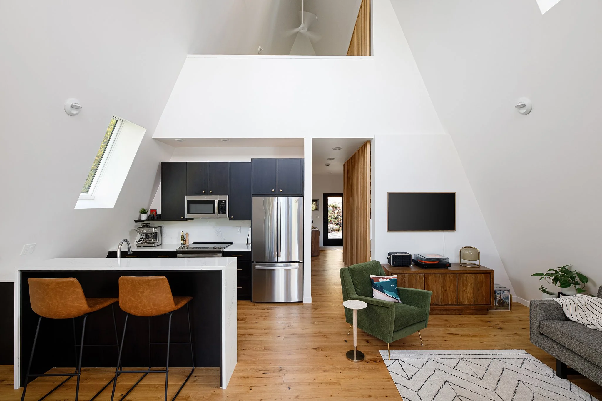 Open-concept living room and kitchen with white walls, wooden floors, a green armchair with a pillow, a gray sofa with a blanket, a wall-mounted TV, and a kitchen area with black cabinets, stainless steel appliances, and a breakfast bar with two brow