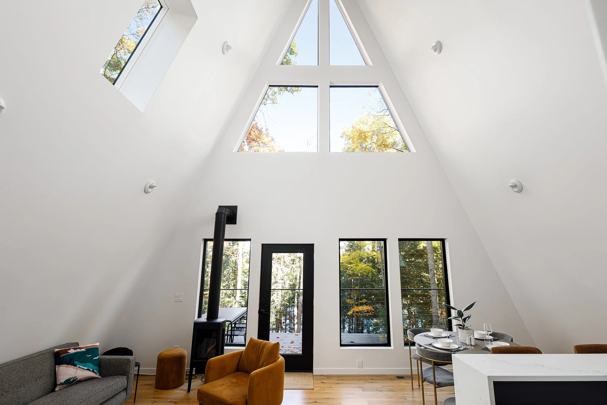 Interior view of a modern living space with high vaulted ceiling, large triangular window, and floor-to-ceiling windows, with furniture and a wood stove.