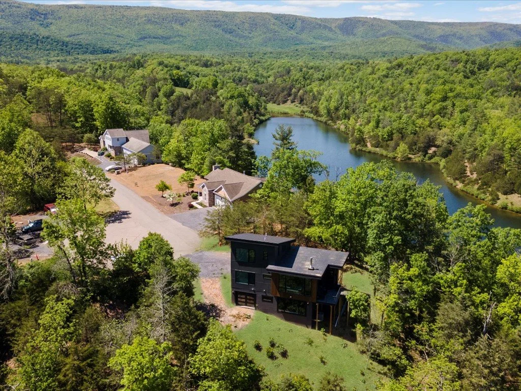  Mountain home designer project featuring an aerial view of a dark-sided home with a raised balcony, wooded hills, and a nearby lake.