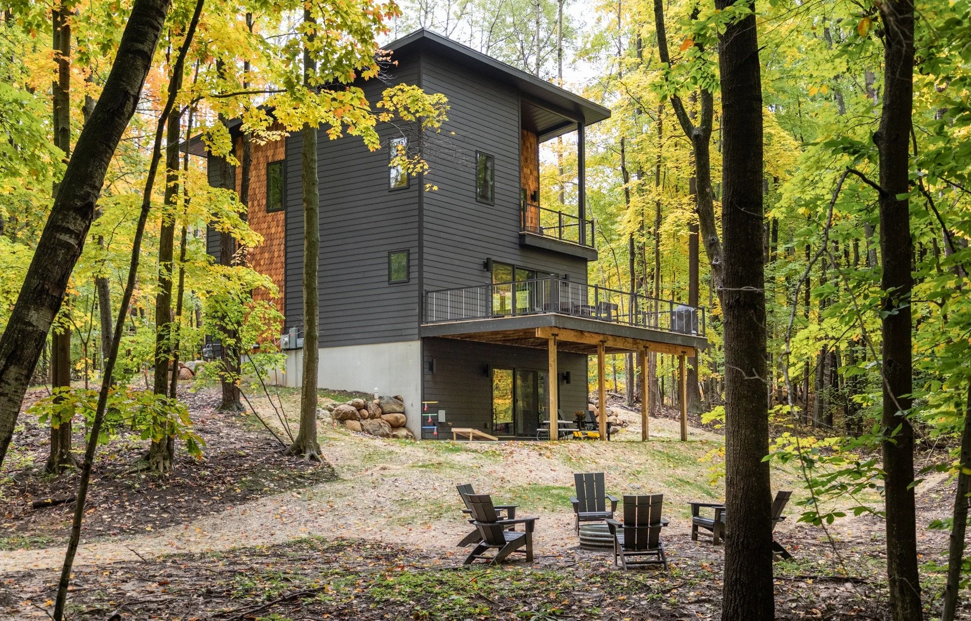 Mountain house architect project featuring dark siding, wood-shingle accents, an upper balcony, and a raised deck above a walkout level in autumn woods.