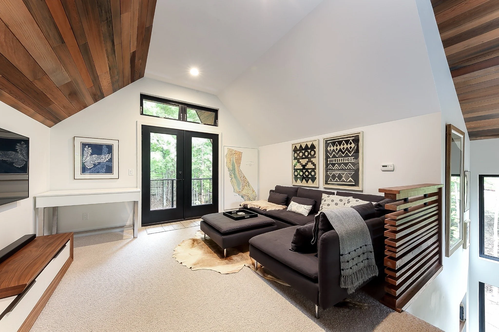  Lake house interior design with a loft sitting area, dark sectional, sloping wood ceiling, and glass doors overlooking trees.
