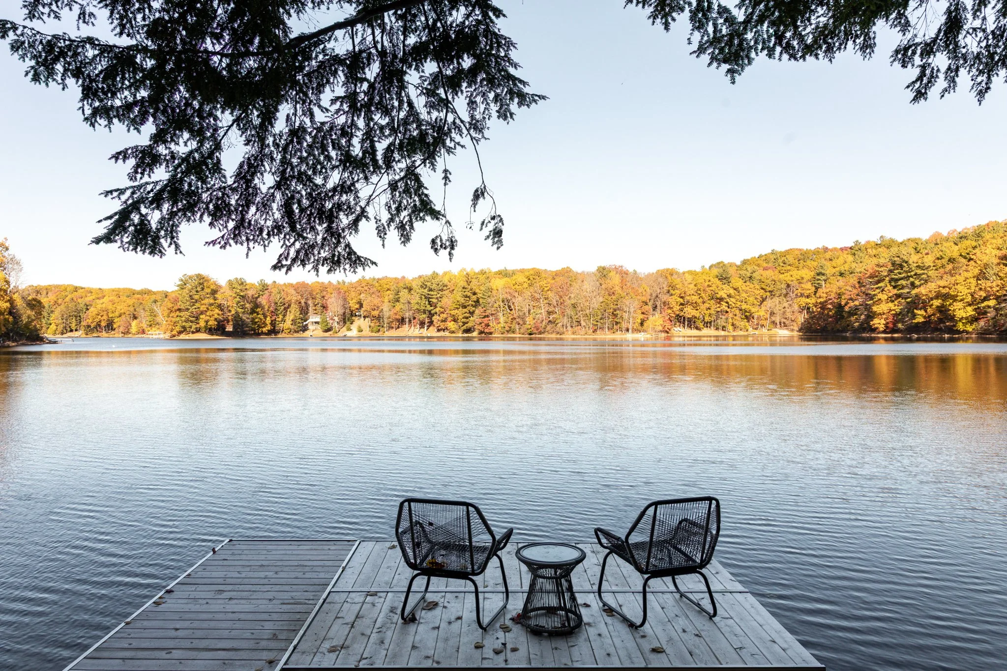 A lakeside view with two black chairs and a small round table on a wooden dock, overlooking calm water surrounded by trees with fall foliage.