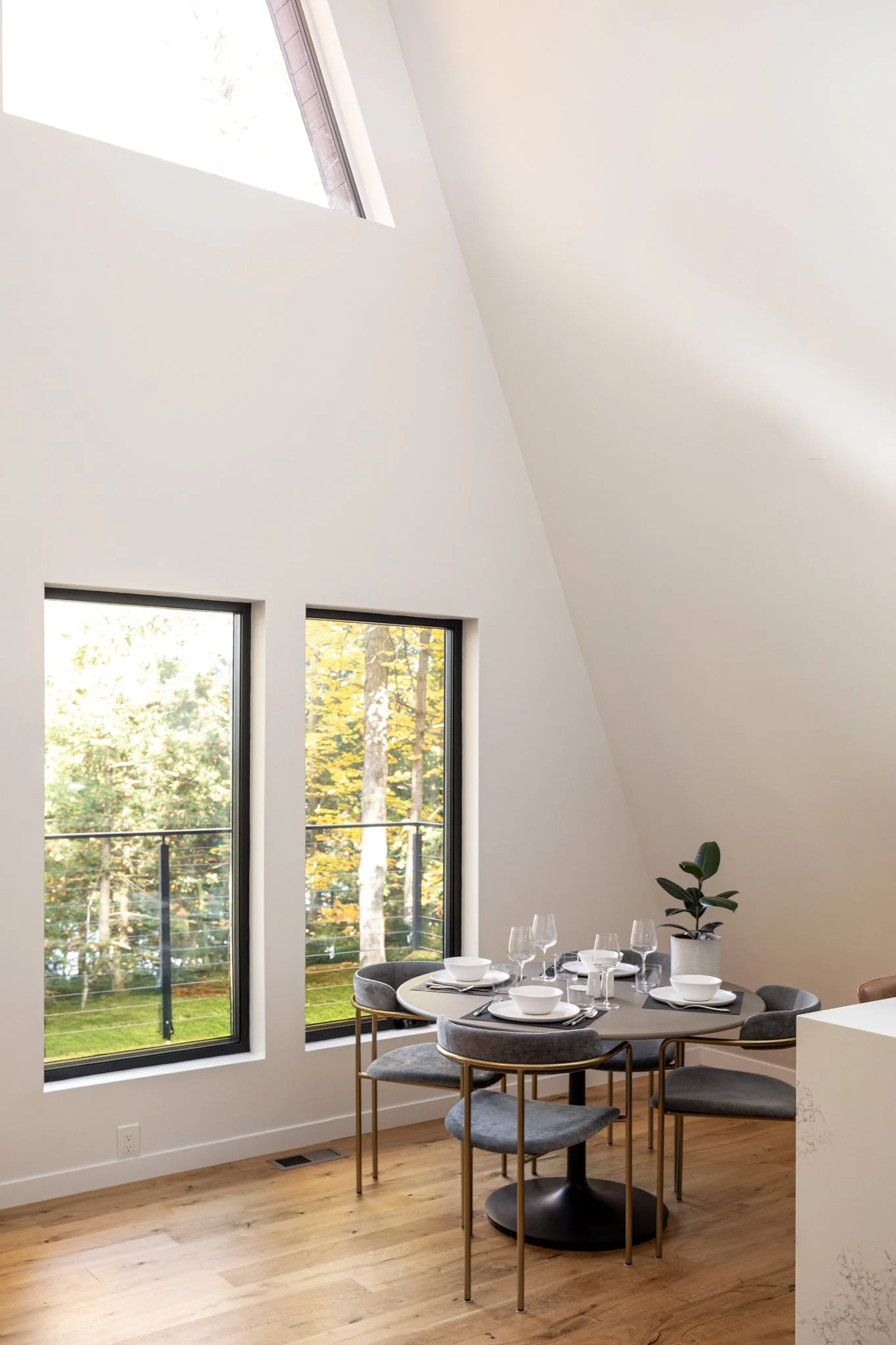 Dining area with four chairs around a round table set with white bowls, glasses, and napkins, next to large windows overlooking a garden, and a potted plant on the table.