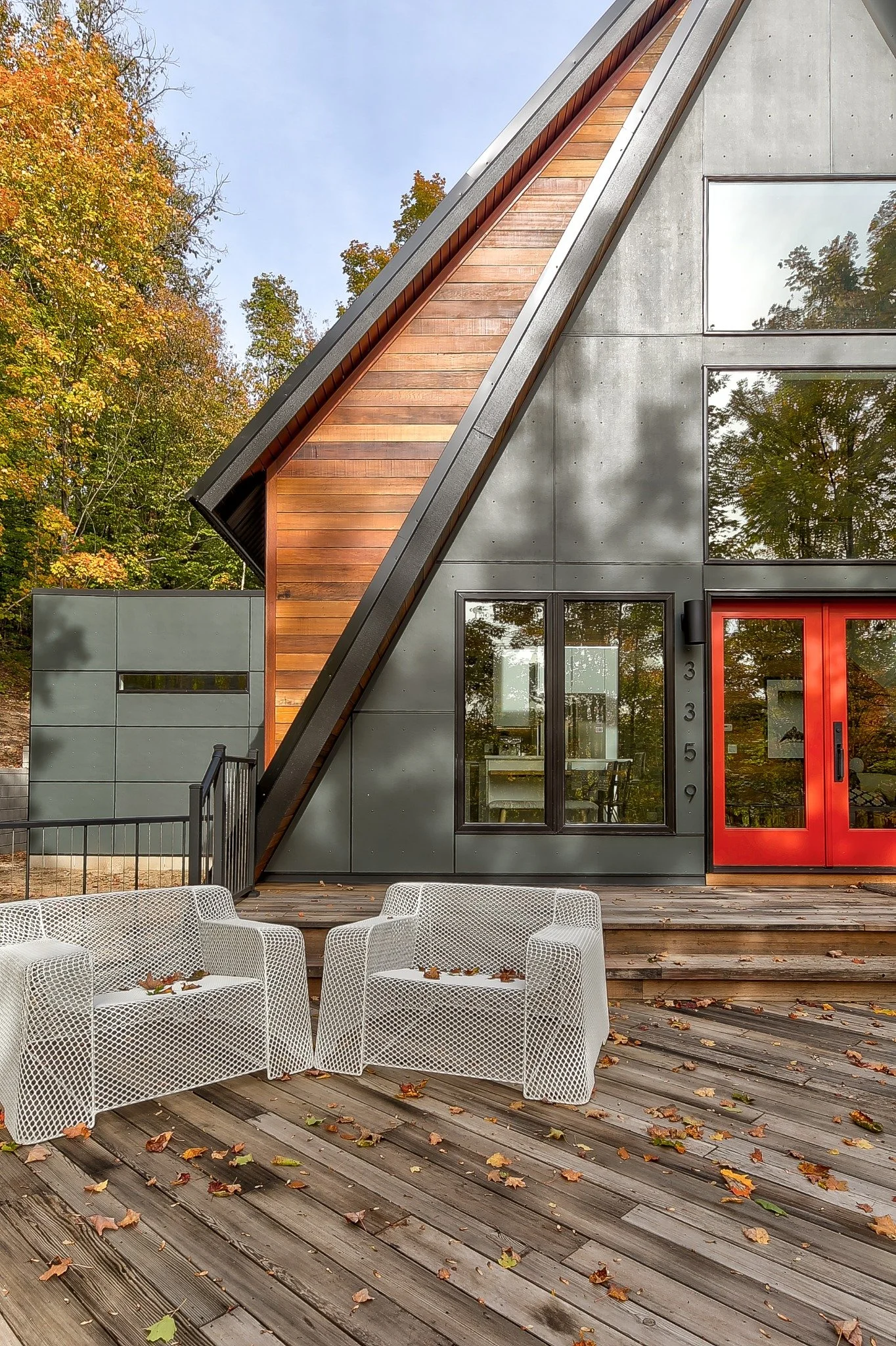 Contemporary lake house design with a steep roof, wood accents, red doors, and a stepped deck among autumn trees.
