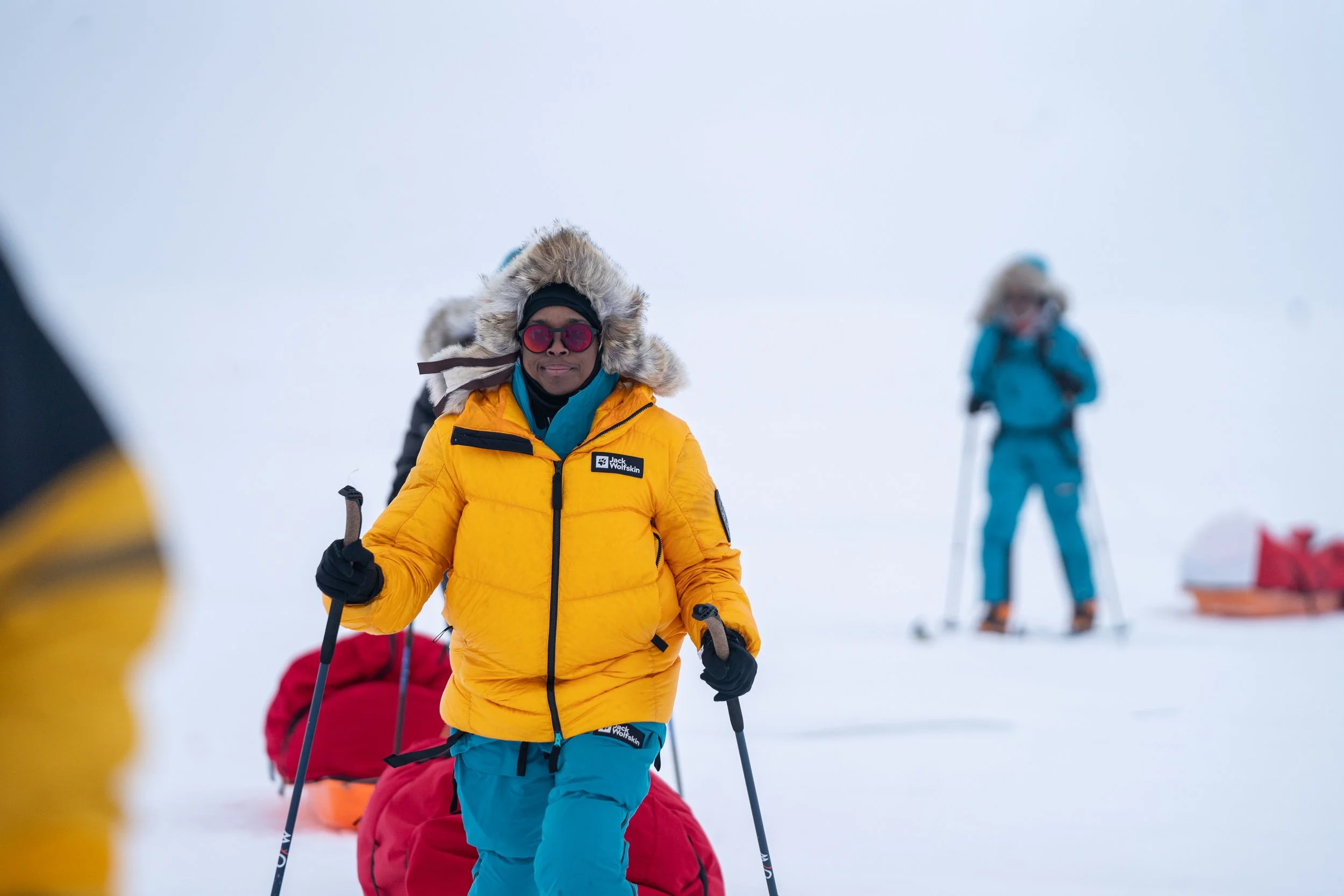 Jeska Clark in a yellow jacket with a fur-lined hood, wearing sunglasses and holding ski poles, standing in a snowy landscape with others in winter gear in the background.