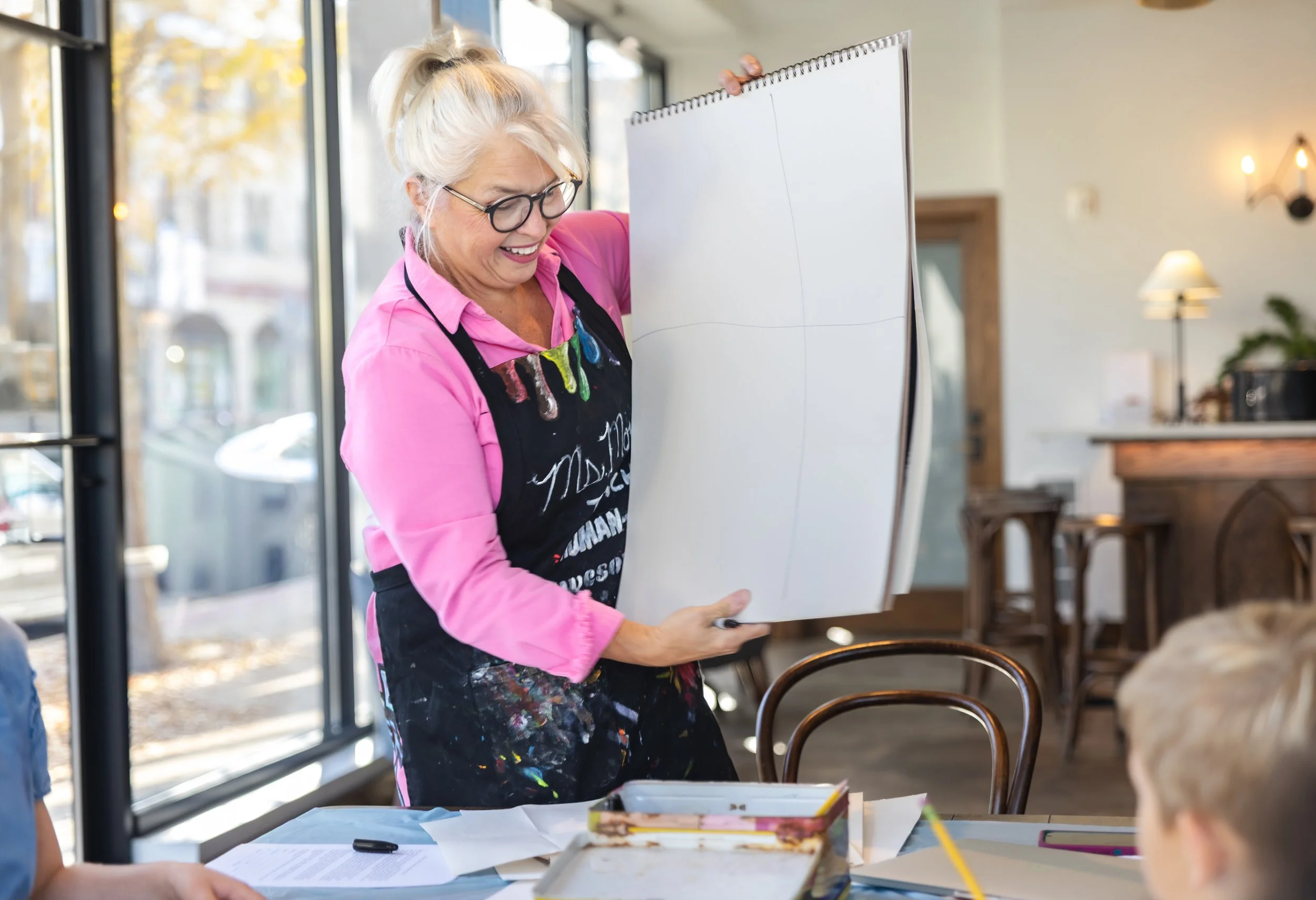 An elderly woman with gray hair, glasses, and a pink shirt, smiling and holding a large white drawing pad, standing inside a bright cafe or restaurant with large windows.
