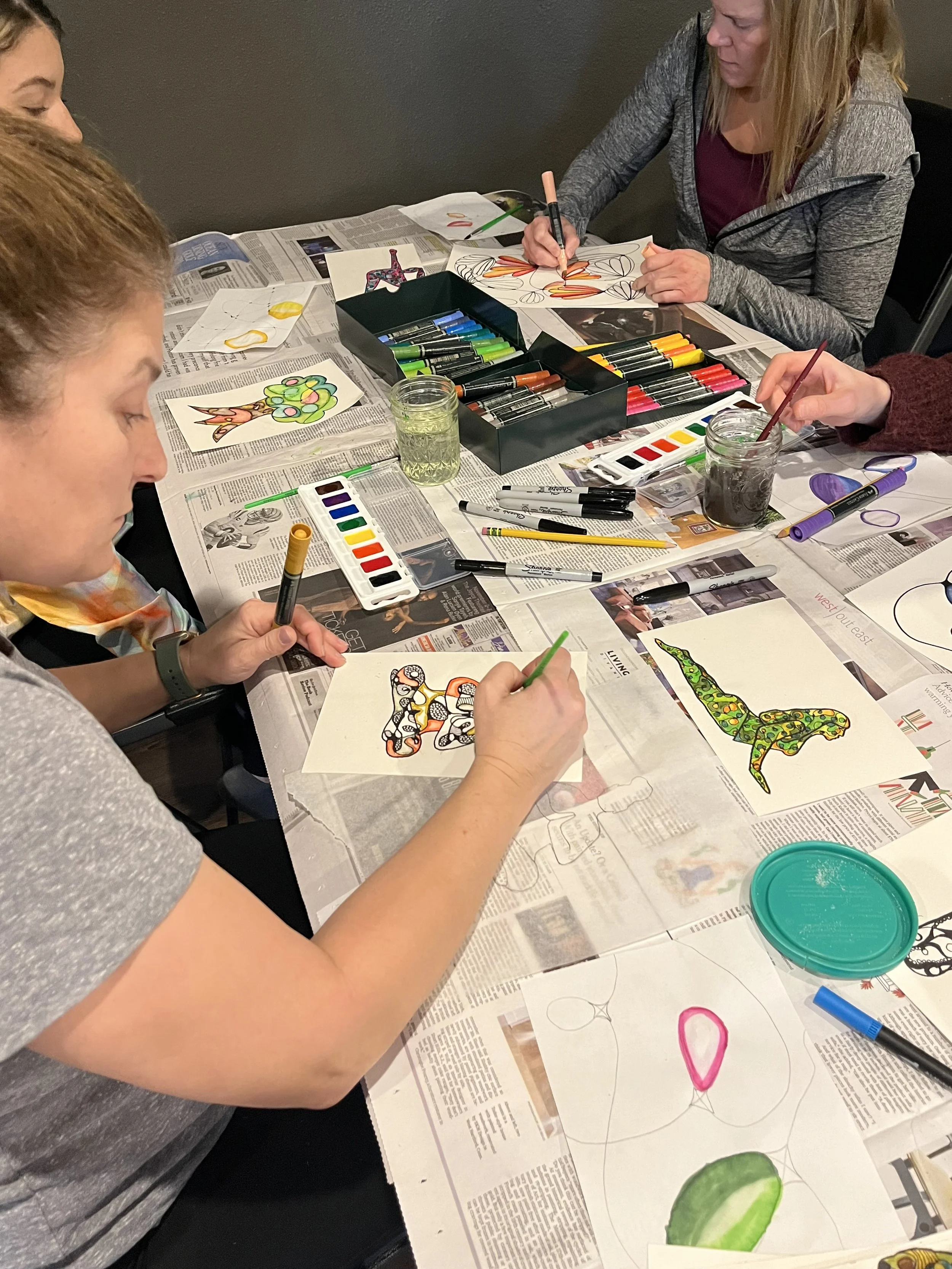 Four women sitting around a table engaged in arts and crafts, painting and drawing with various markers and paints, with colorful paper art projects and newspaper covering the table.