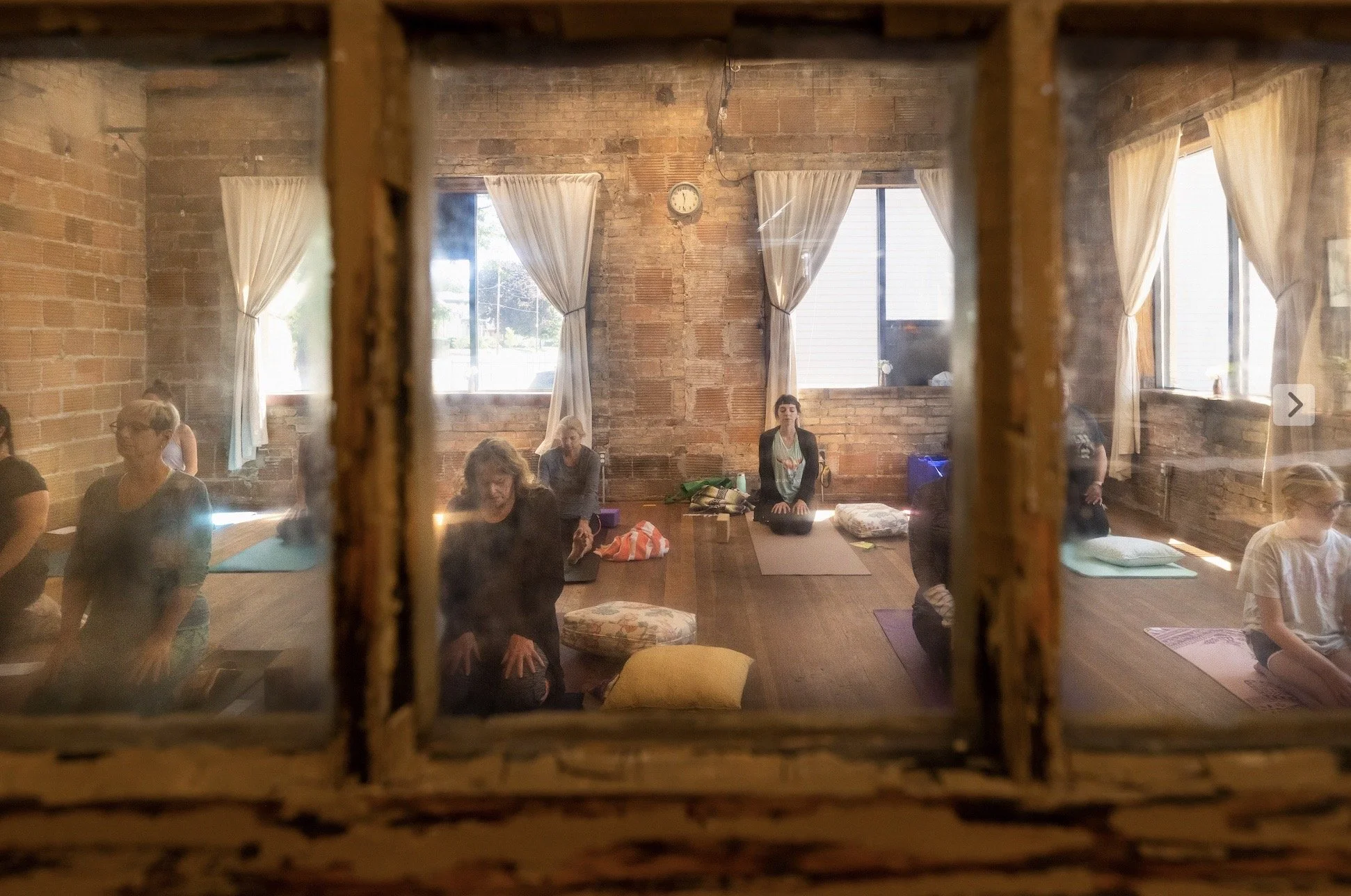 People participating in a yoga class in a brick-walled studio viewed through a window with divided panes.