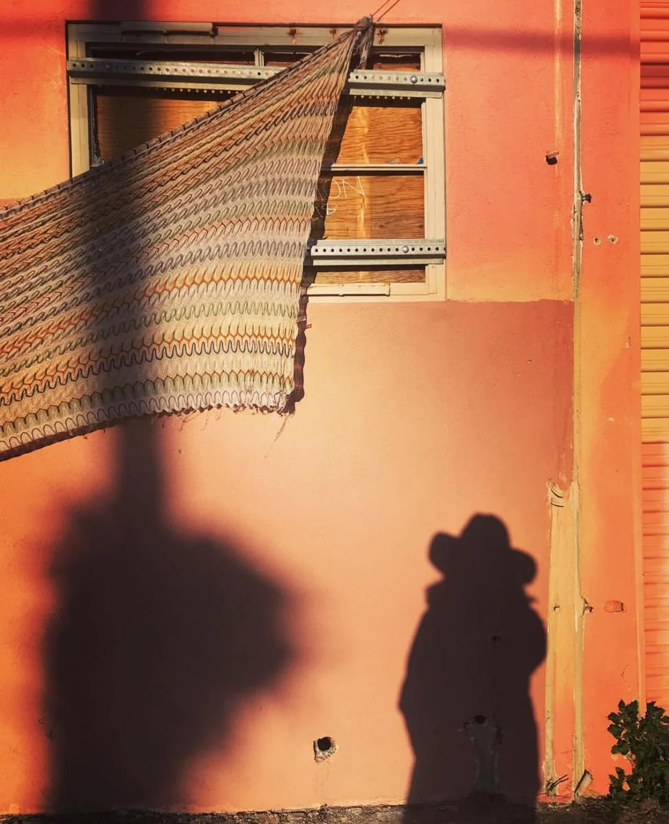 A building with a pink exterior wall, a shadow of a person wearing a hat cast onto the wall, and a window with wooden boards covering the glass. A decorative fabric is hanging outside the window.