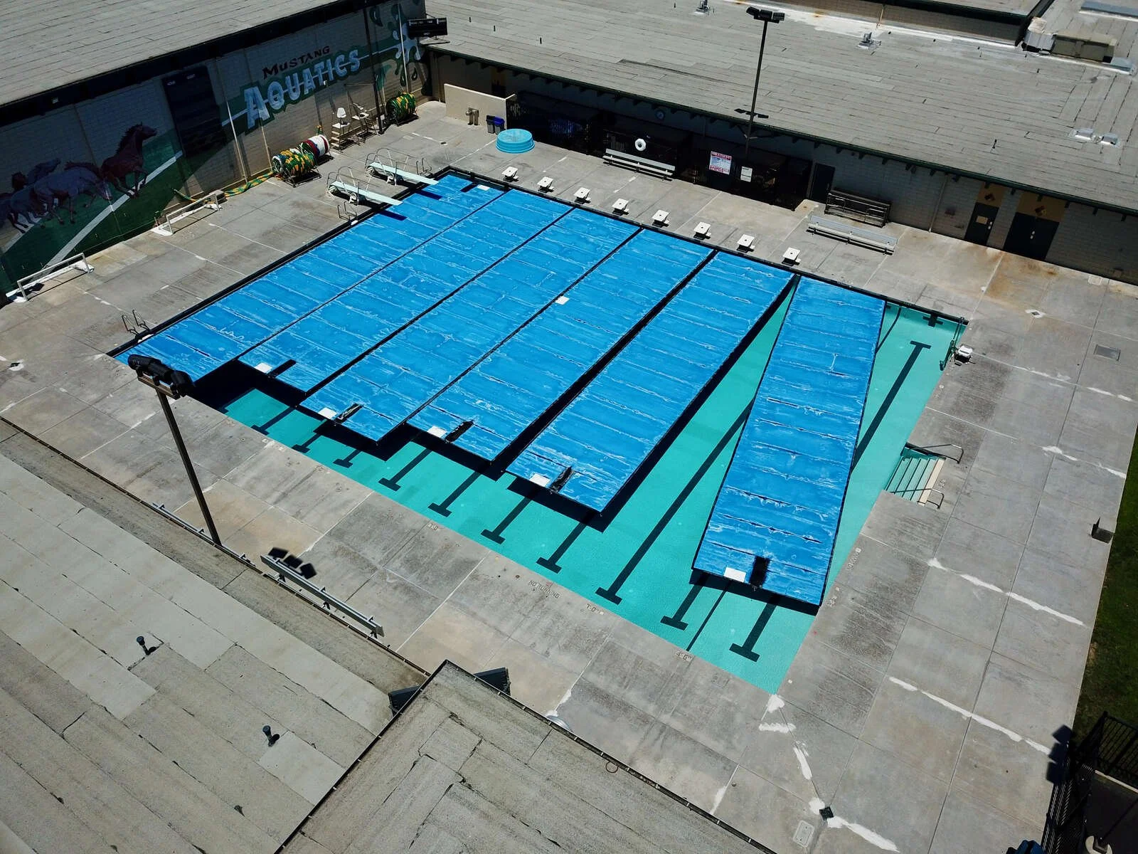 A swimming pool covered with a blue tarp, located in a large open concrete area outside a building with the sign 'Mustang Aquatics', seen from an aerial view.