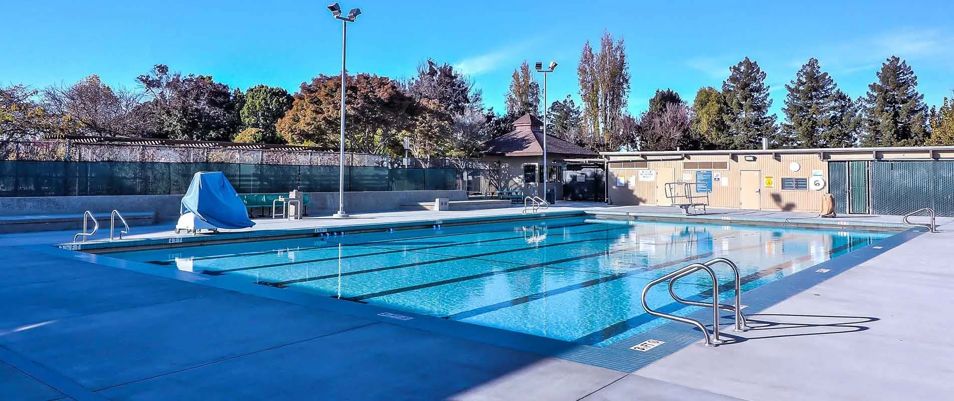 Empty outdoor swimming pool with blue water, surrounded by concrete deck, with trees and a small building in the background.