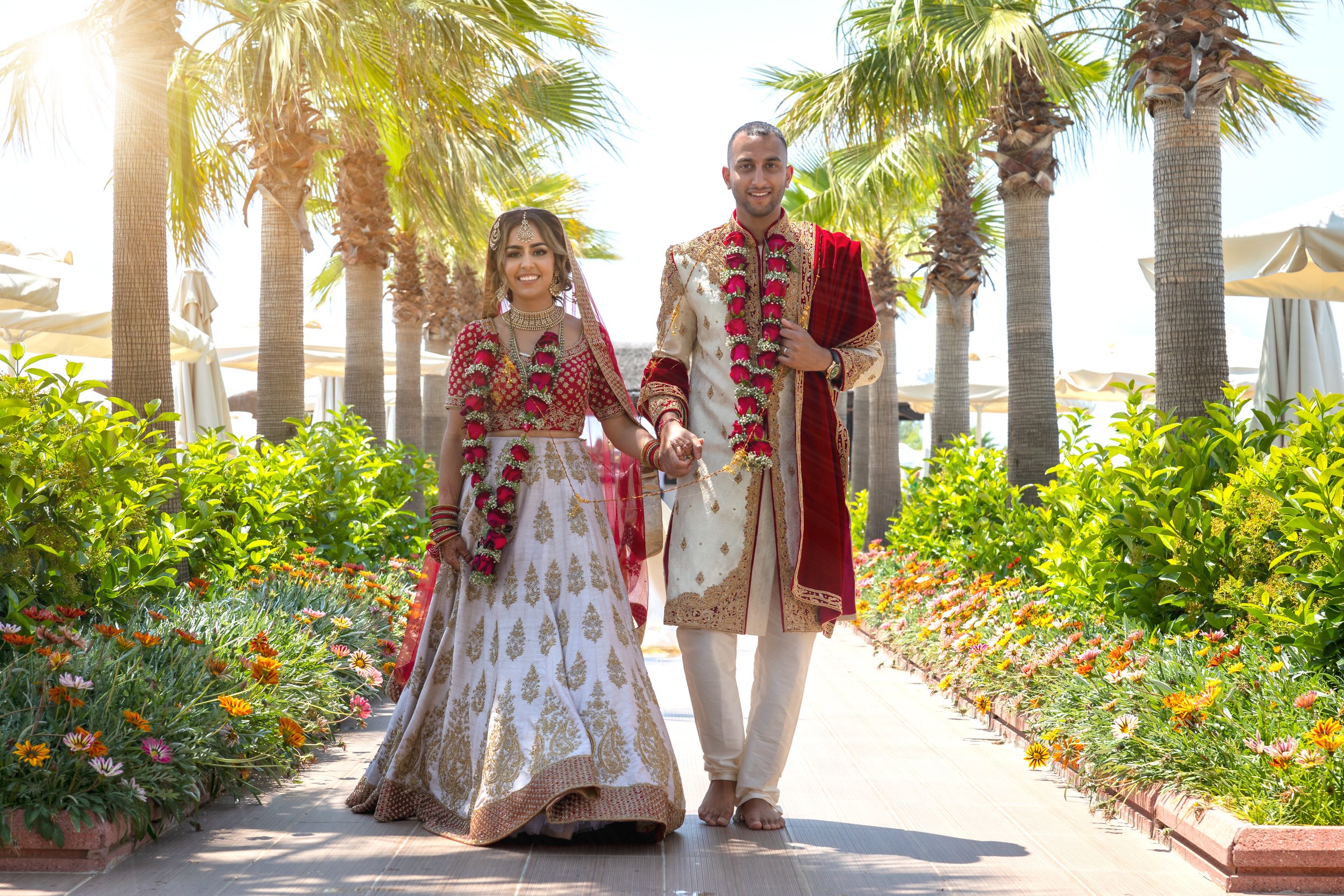 A bride and groom portrait taken at their wedding in Turkey, wearing tradition Indian wedding outfits. The couple are walking down a path surrounded by palm trees.