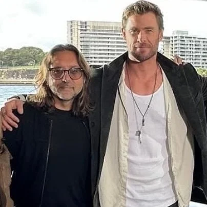 Two men standing outdoors with city buildings in the background, one with glasses and long hair, the other with short hair and a beard, both casually dressed.