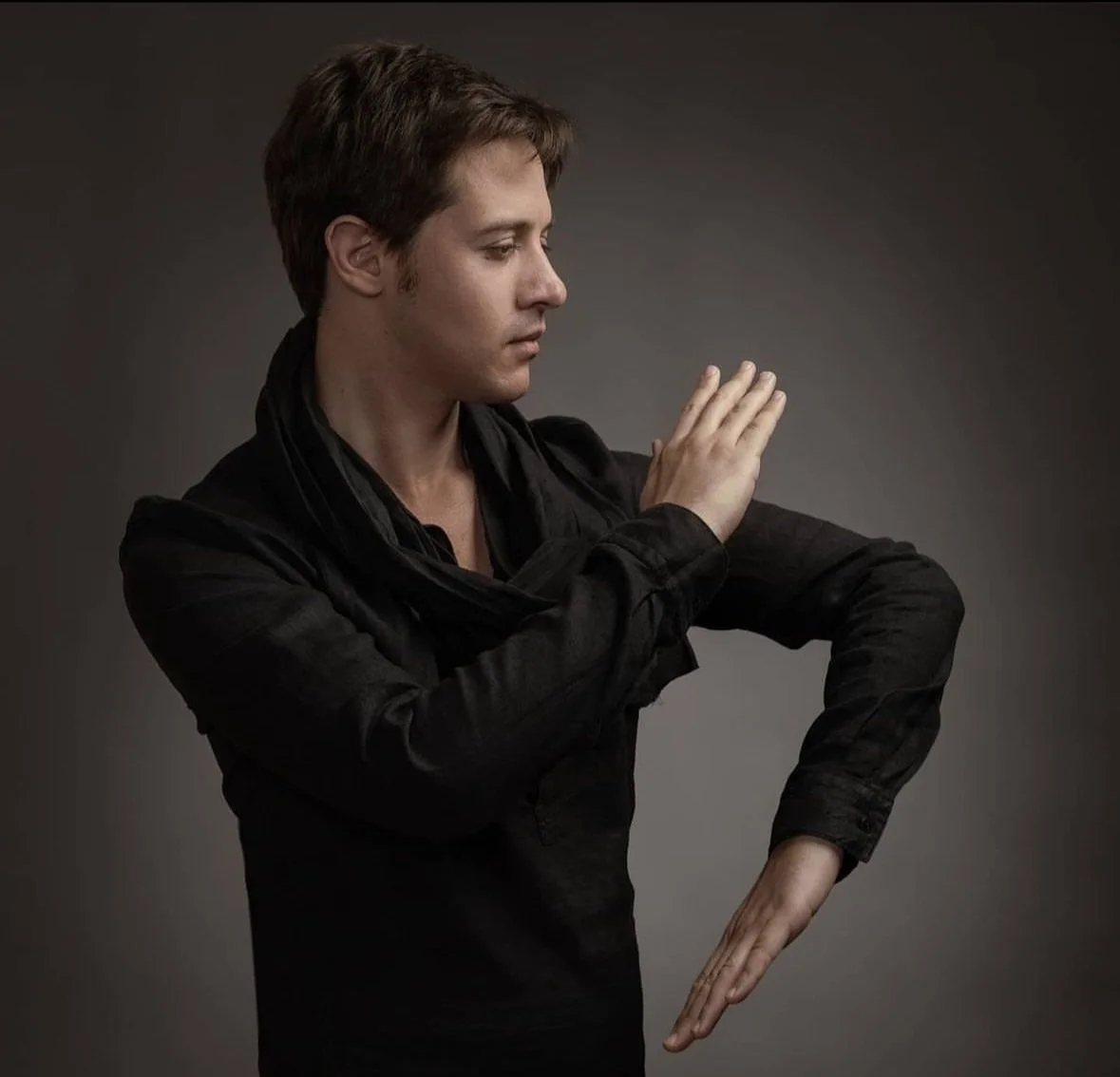 A man with brown hair, wearing a black shirt and scarf, holds his hands in a martial arts pose against a dark gray background.