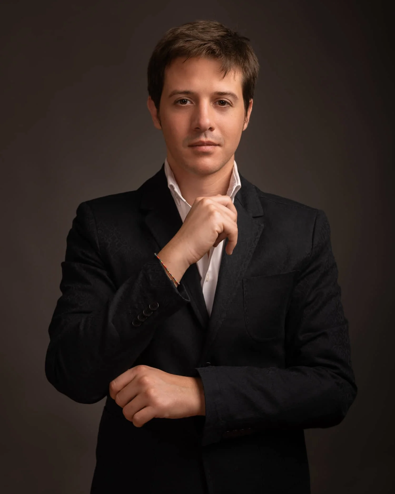 A young man with dark hair wearing a black blazer over a white collared shirt, posing against a dark background, with one hand resting near his chin and the other crossed over his stomach.