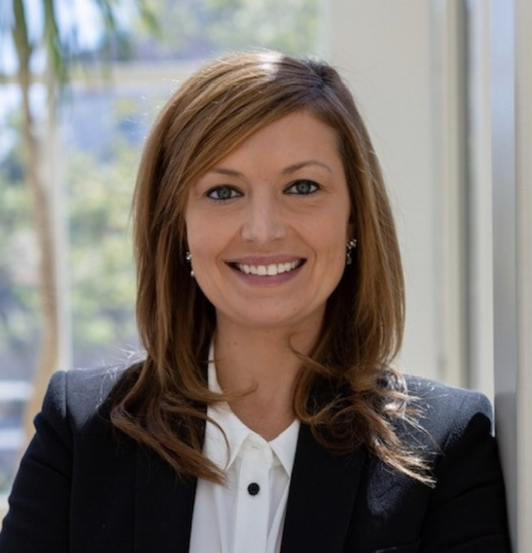 A professional woman with shoulder-length brown hair, smiling, wearing a dark blazer and white shirt, sitting indoors near a window with a blurred outdoor background.