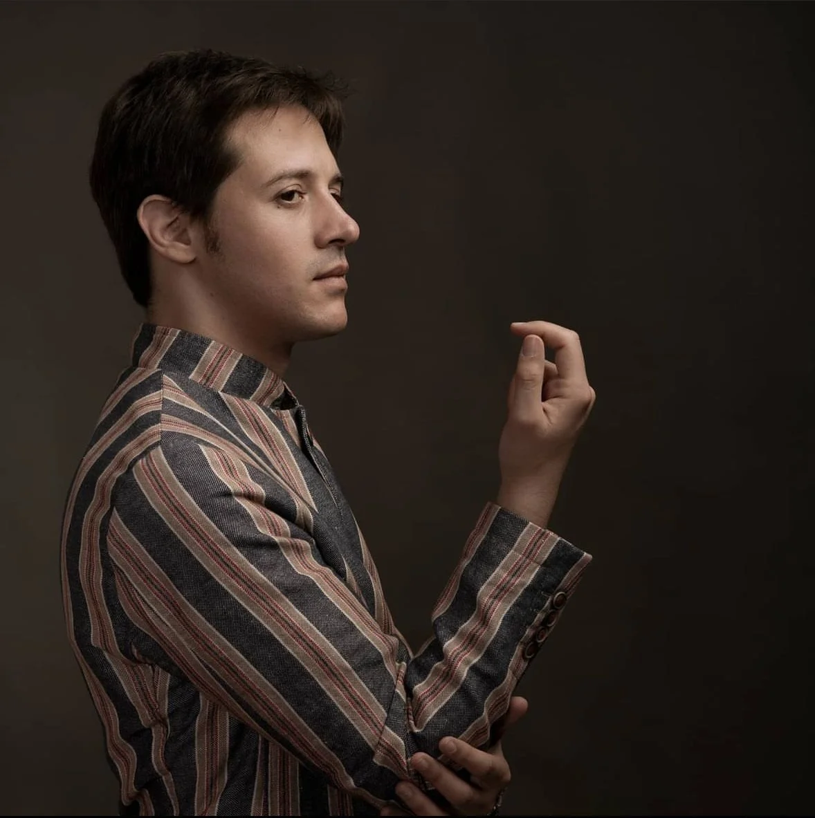 Side profile of a man with brown hair wearing a striped blazer against a dark background.