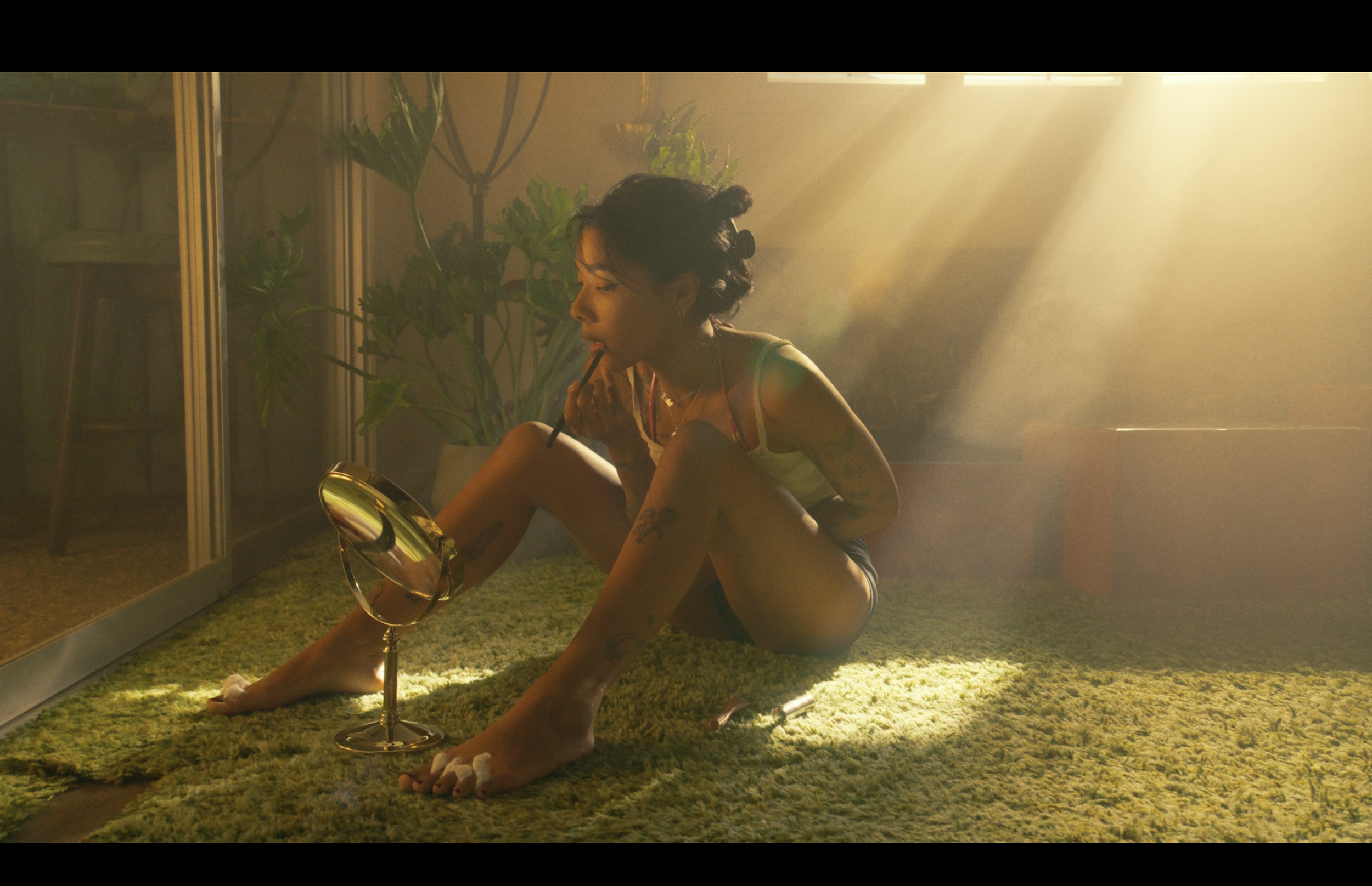 A woman with short dark hair styling her hair while sitting on a green shag rug in a room illuminated with warm sunlight, surrounded by plants and a large mirror.