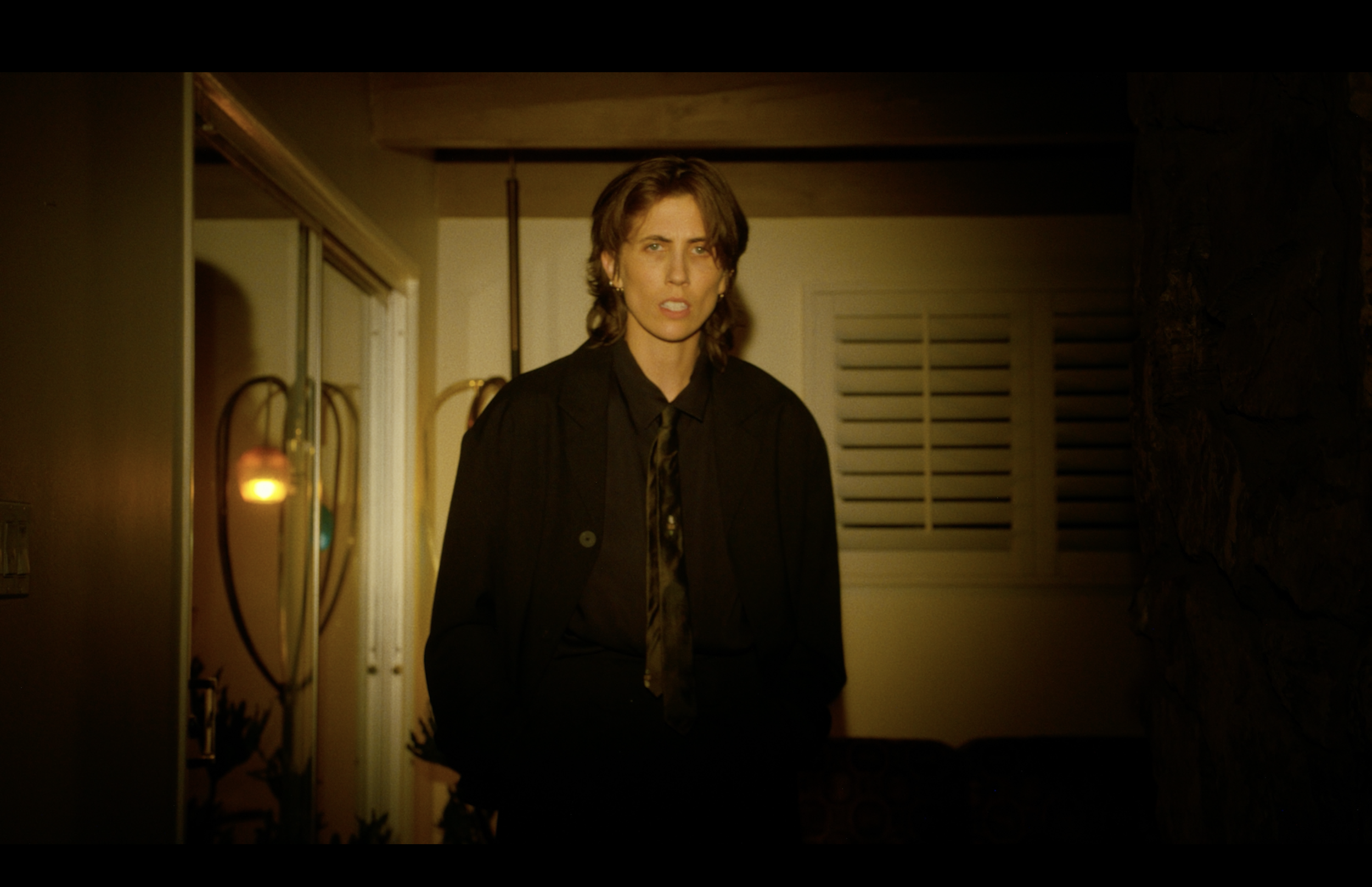 Woman with short brown hair wearing a black suit and tie, standing indoors in a dimly lit room.