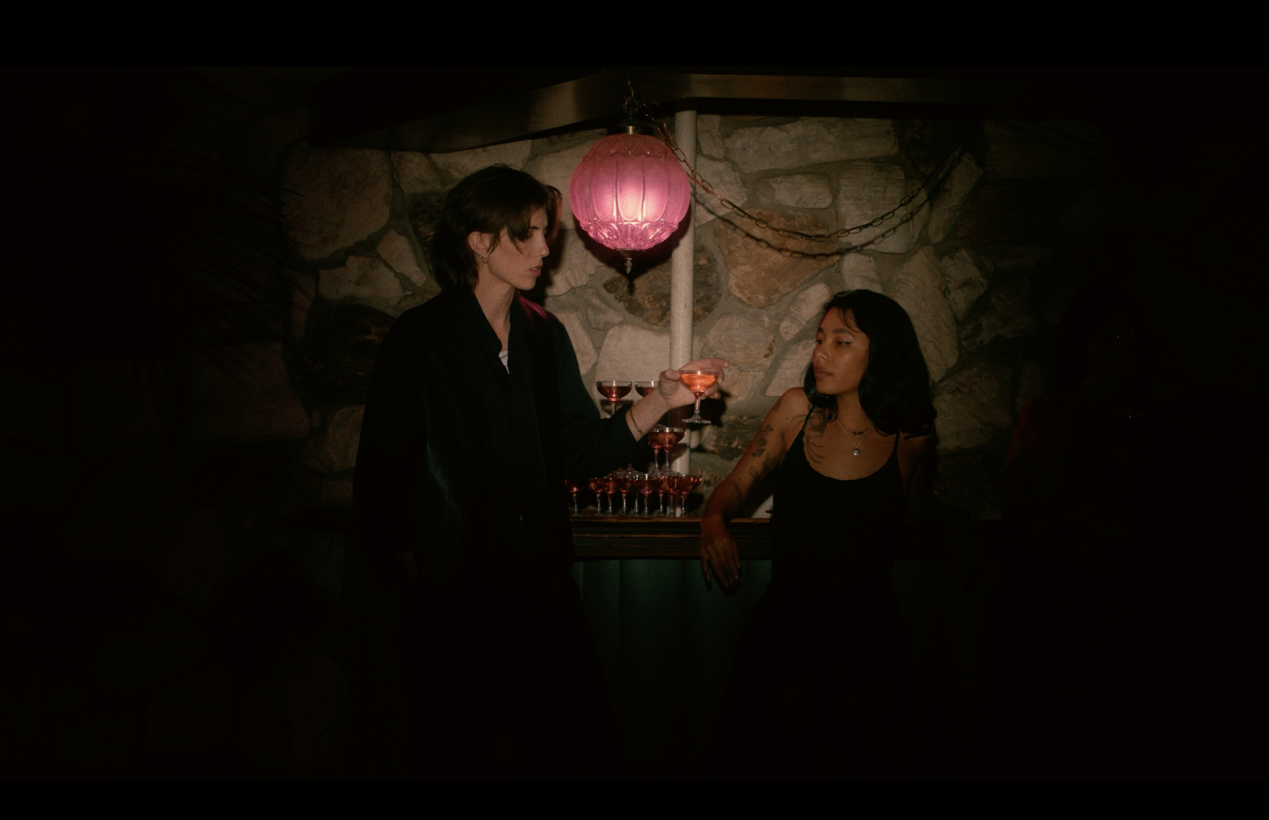 Two women in a dimly lit room with a stone wall, one woman with short brown hair holding a drink, and the other with long dark hair sitting at a table.
