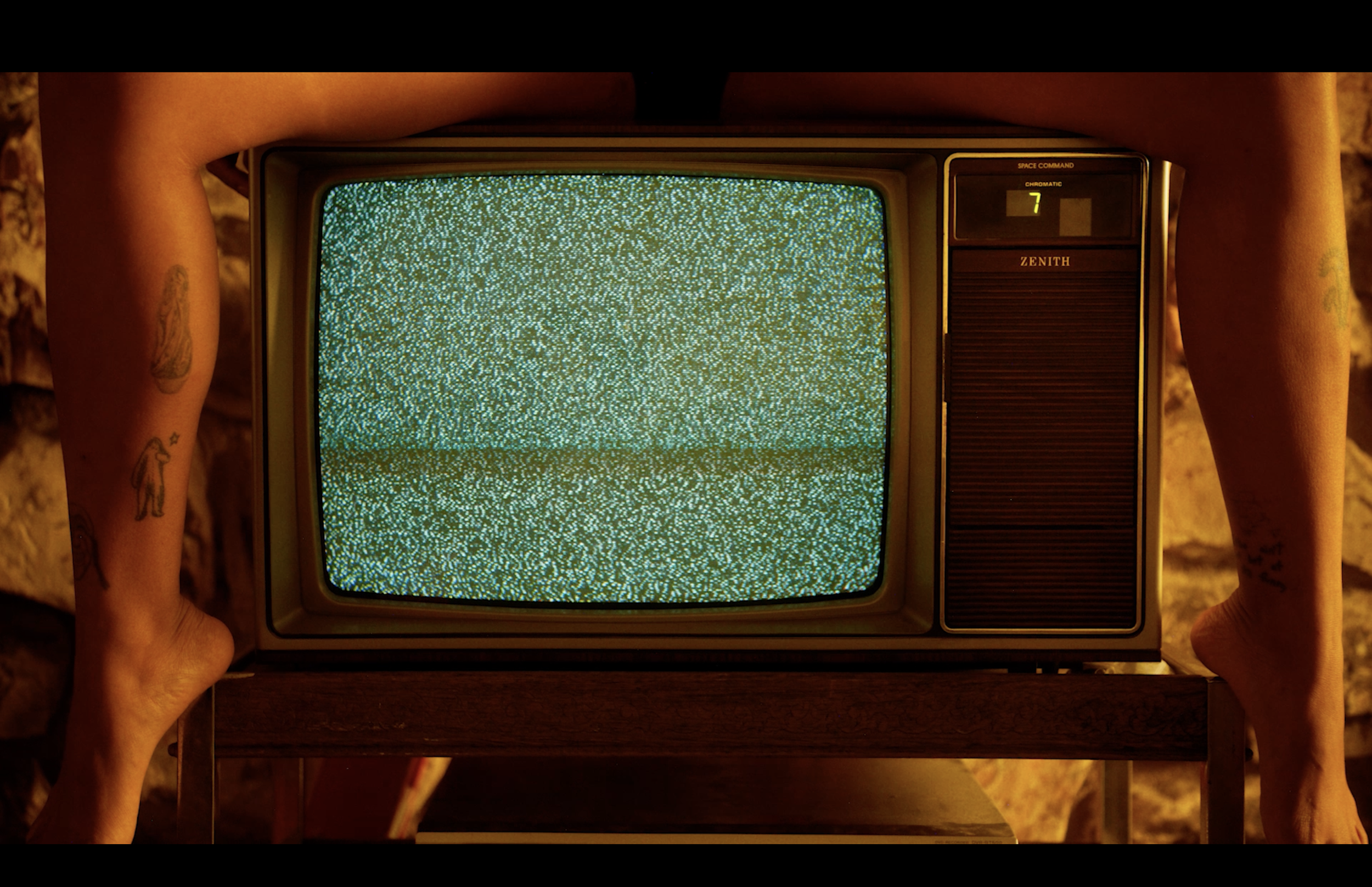 Person holding an old television with a static screen, standing on a wooden surface, tattoos visible on their arms.