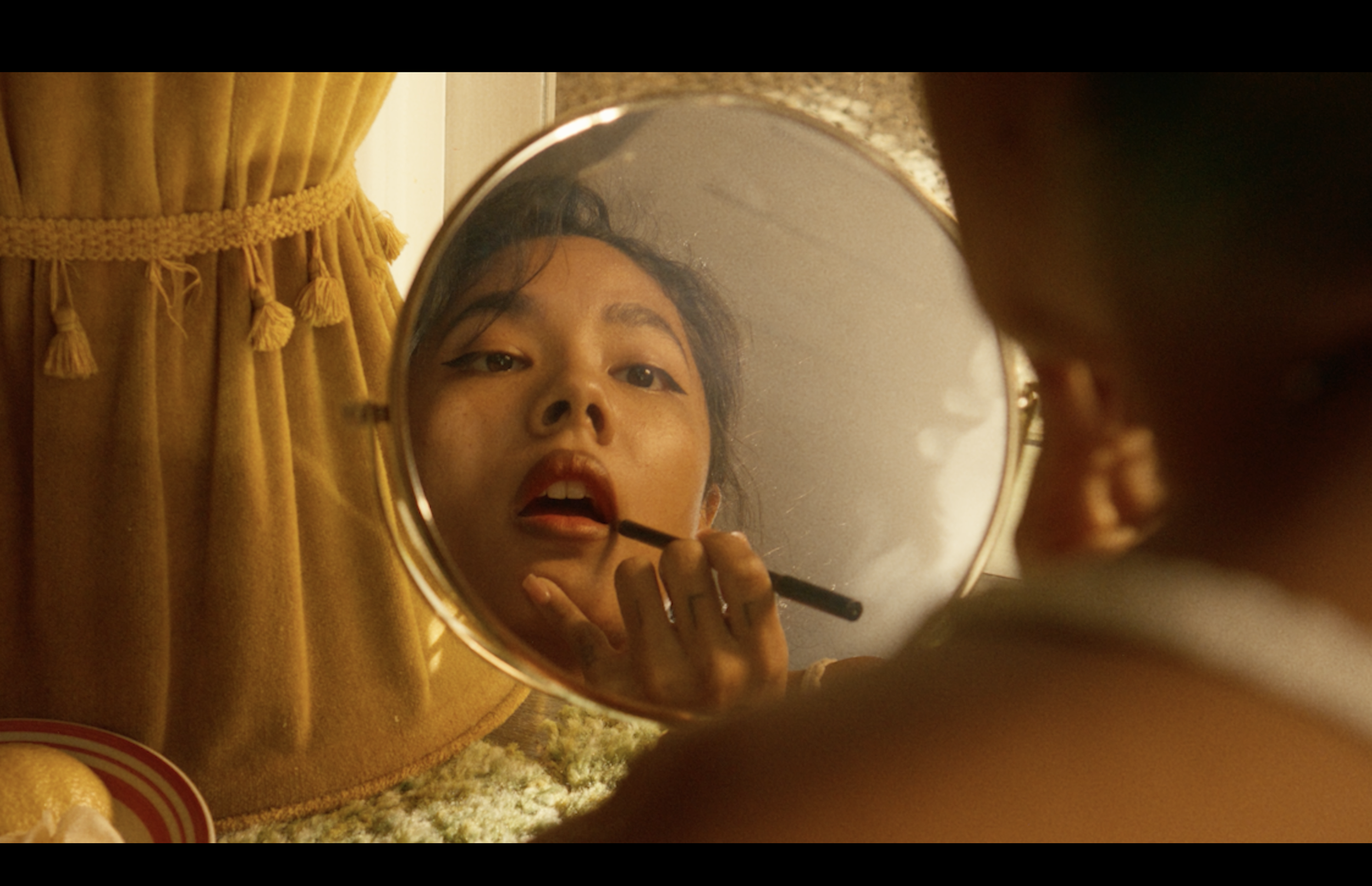 A woman applies lipstick while looking into a handheld mirror on a kitchen countertop with a yellow curtain in the background.