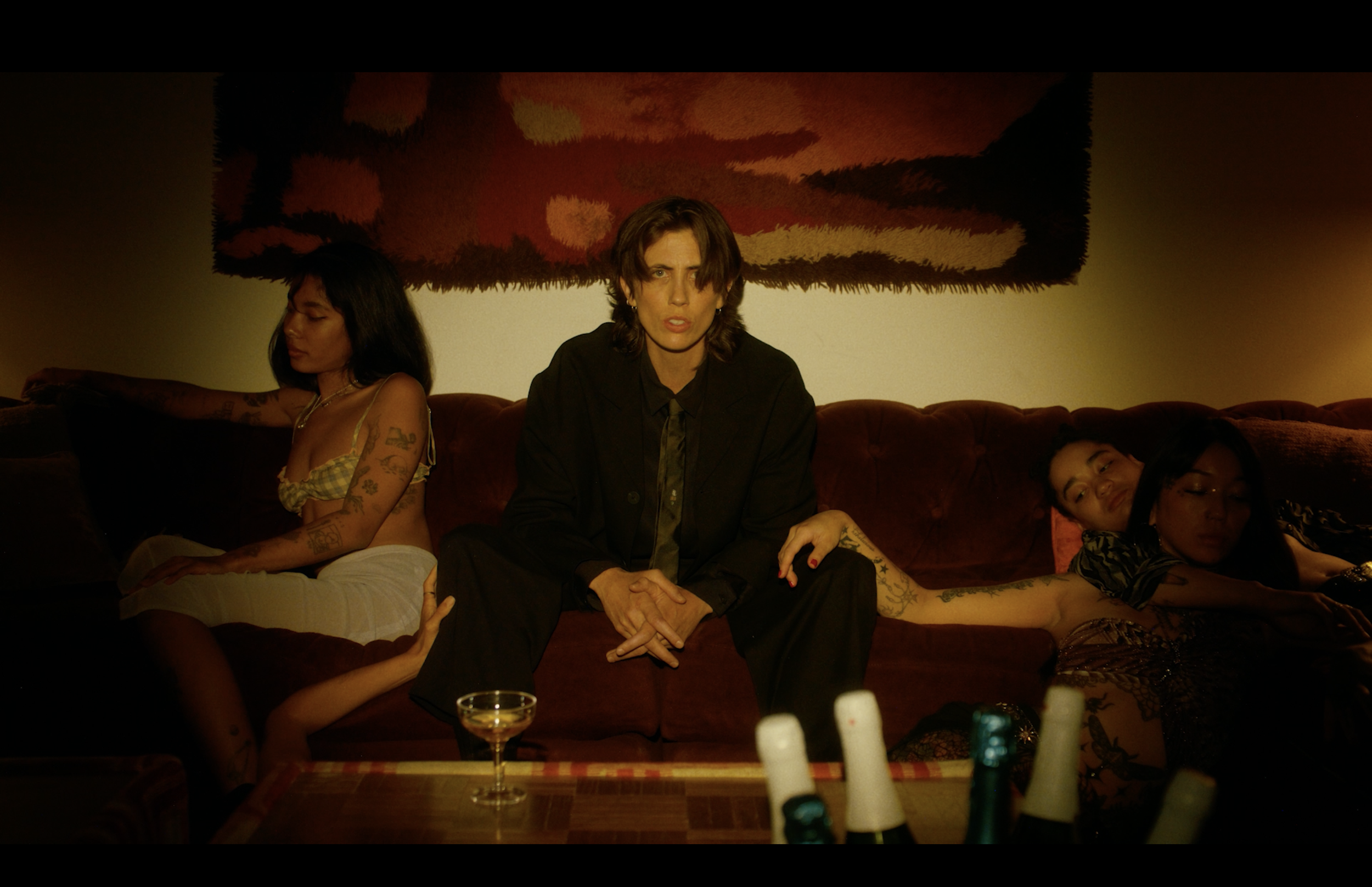 A woman sitting on a red couch with three women lying beside her, in a dimly lit room with a colorful abstract wall hanging.