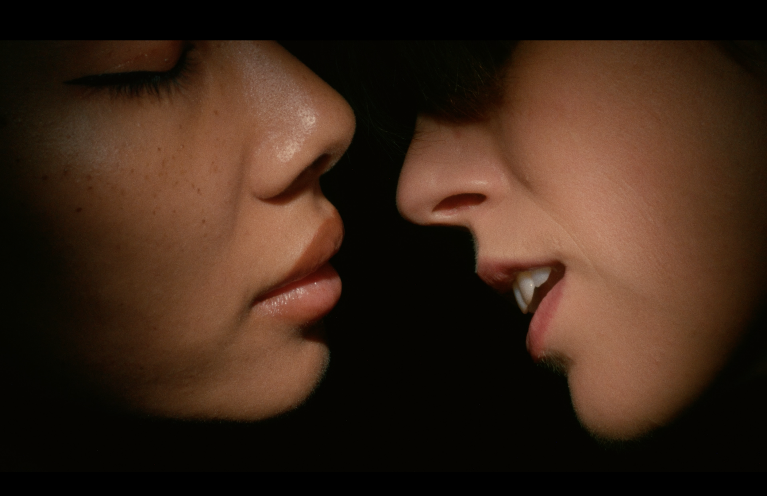 Two women are close to each other, nose to nose, with one woman smiling showing teeth and the other with closed eyes.