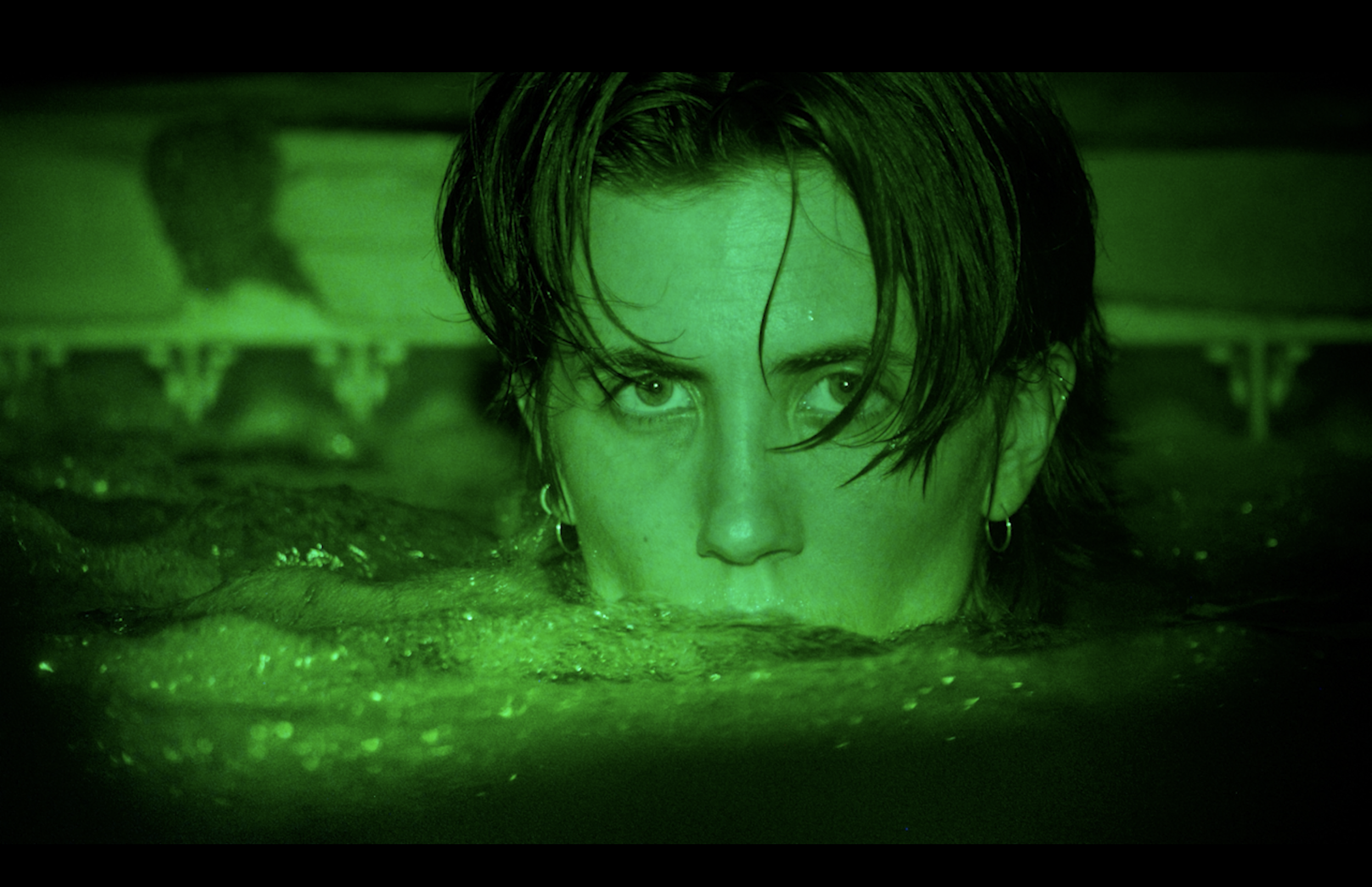 A person with wet dark hair and earrings is submerged up to their nose in water, illuminated with green light, with their eyes staring ahead.