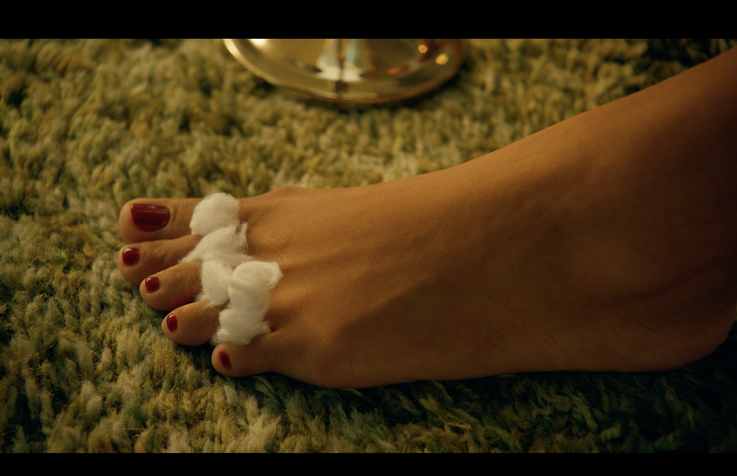 A hand with red nail polish resting on a multicolored textured carpet, with a white fuzzy decorative item around the wrist and a shiny silver object in the background.