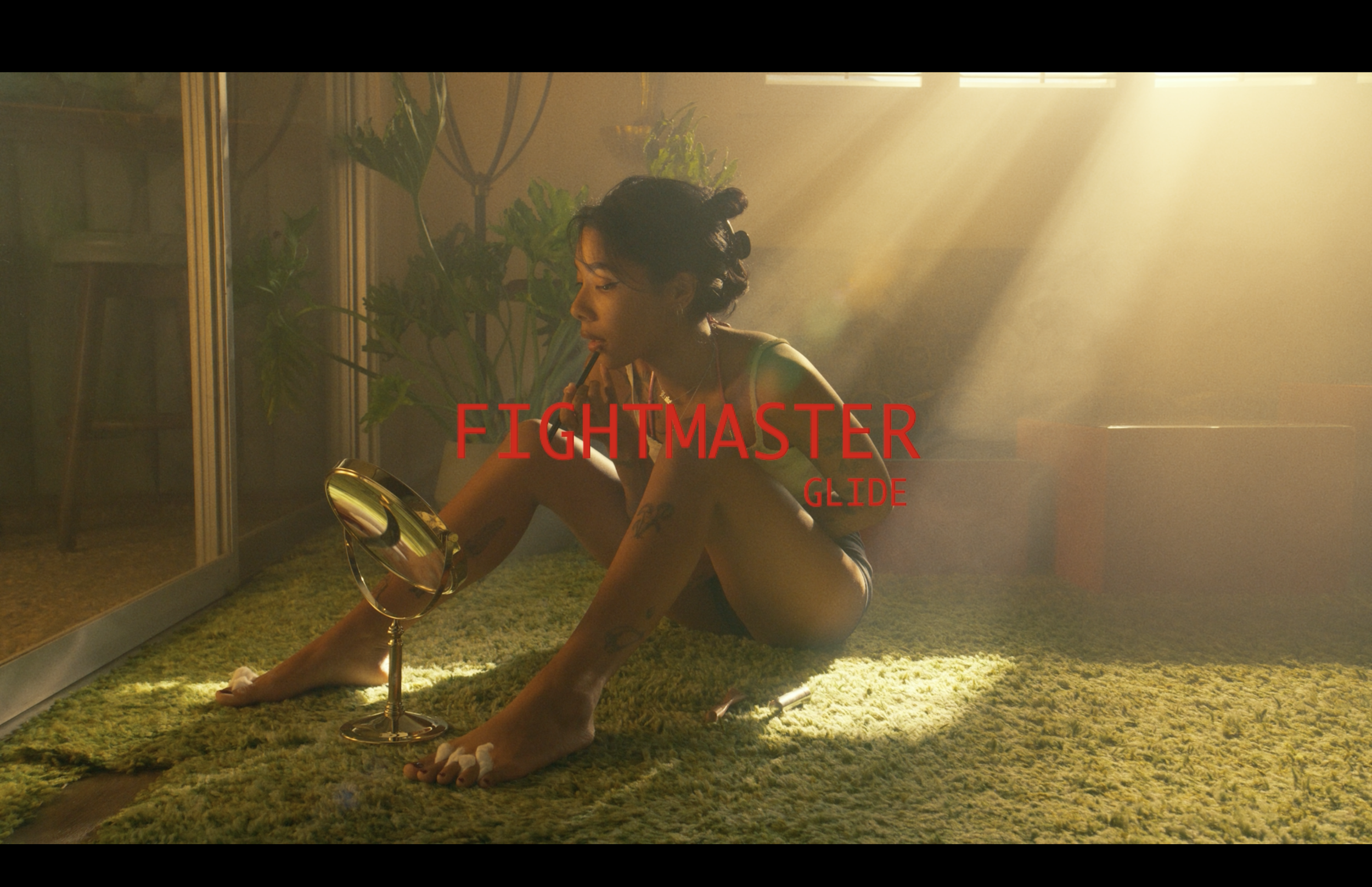 A woman with tattoos and dark curly hair sits on a green shaggy carpet, applying lipstick while looking into a mirror. She is near a glass door with plants inside, illuminated by warm sunlight, with a bottle and makeup items around her. The text 'FIG