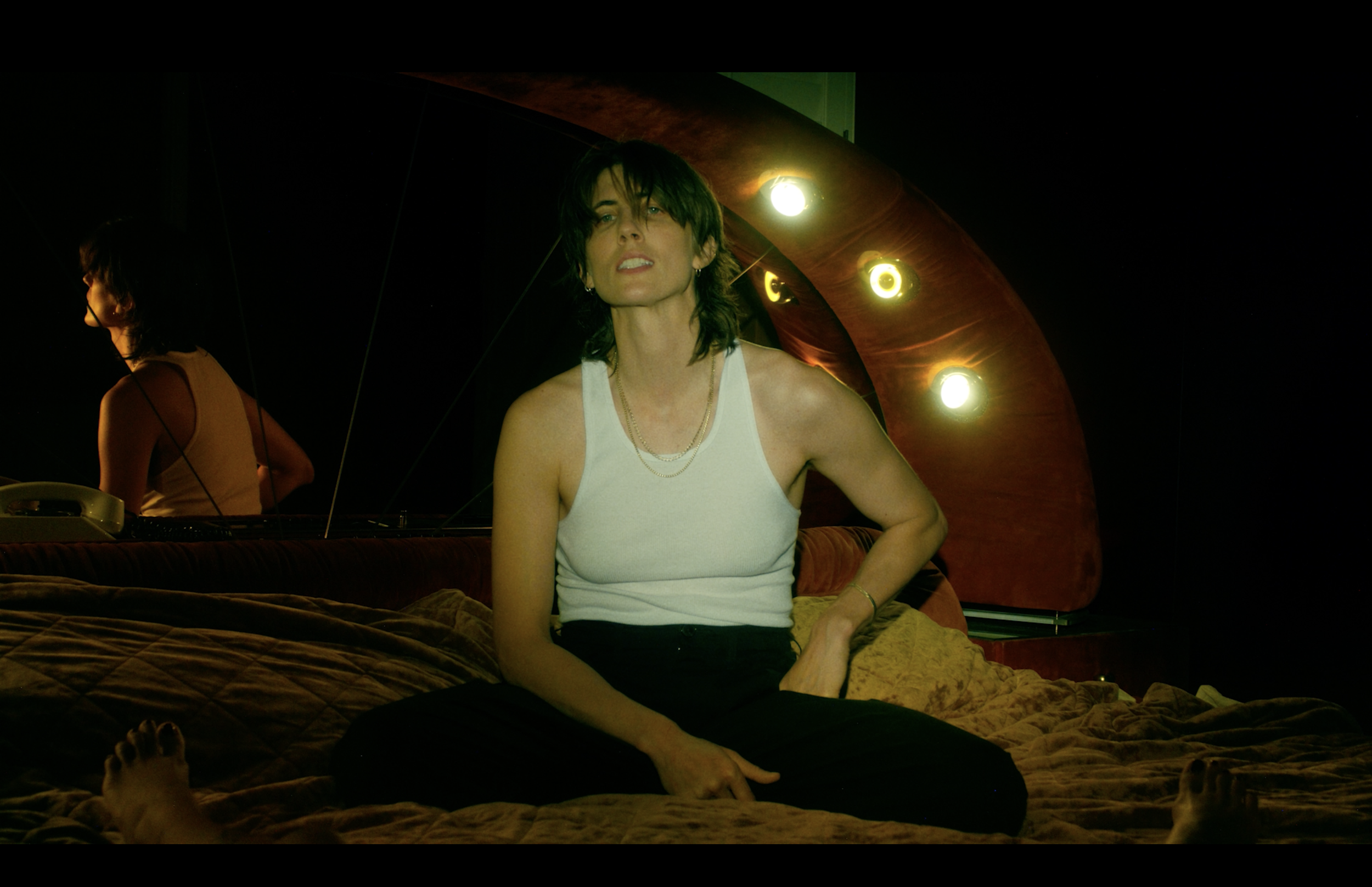 A woman with shoulder-length dark hair wearing a white tank top sitting on a bed with a dark quilt. She is in front of a mirror that reflects her back and part of the room. The room has dark walls, with warm lighting and a row of exposed light bulbs 