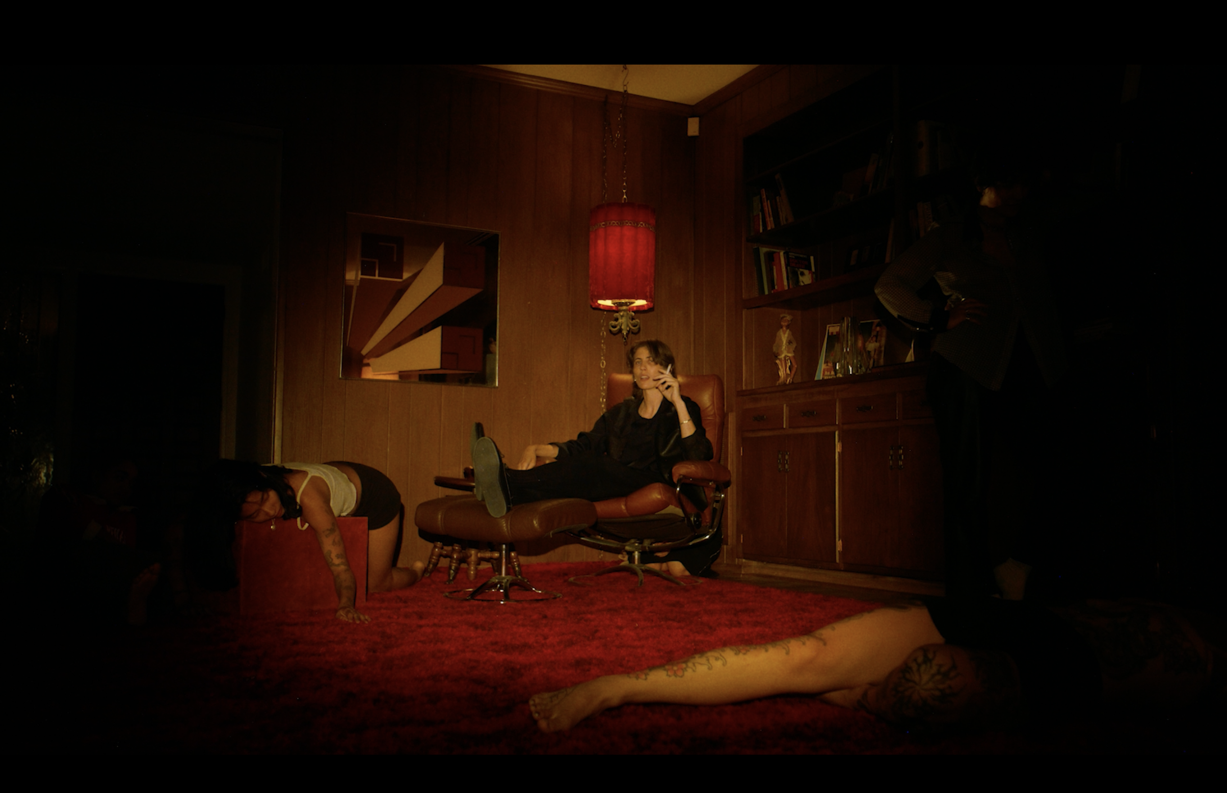 A woman sitting in a leather chair while talking on a phone, in a dimly lit room with wood-paneled walls and bookshelves, and a red hanging lamp. There are people on the floor and a person standing to the right.