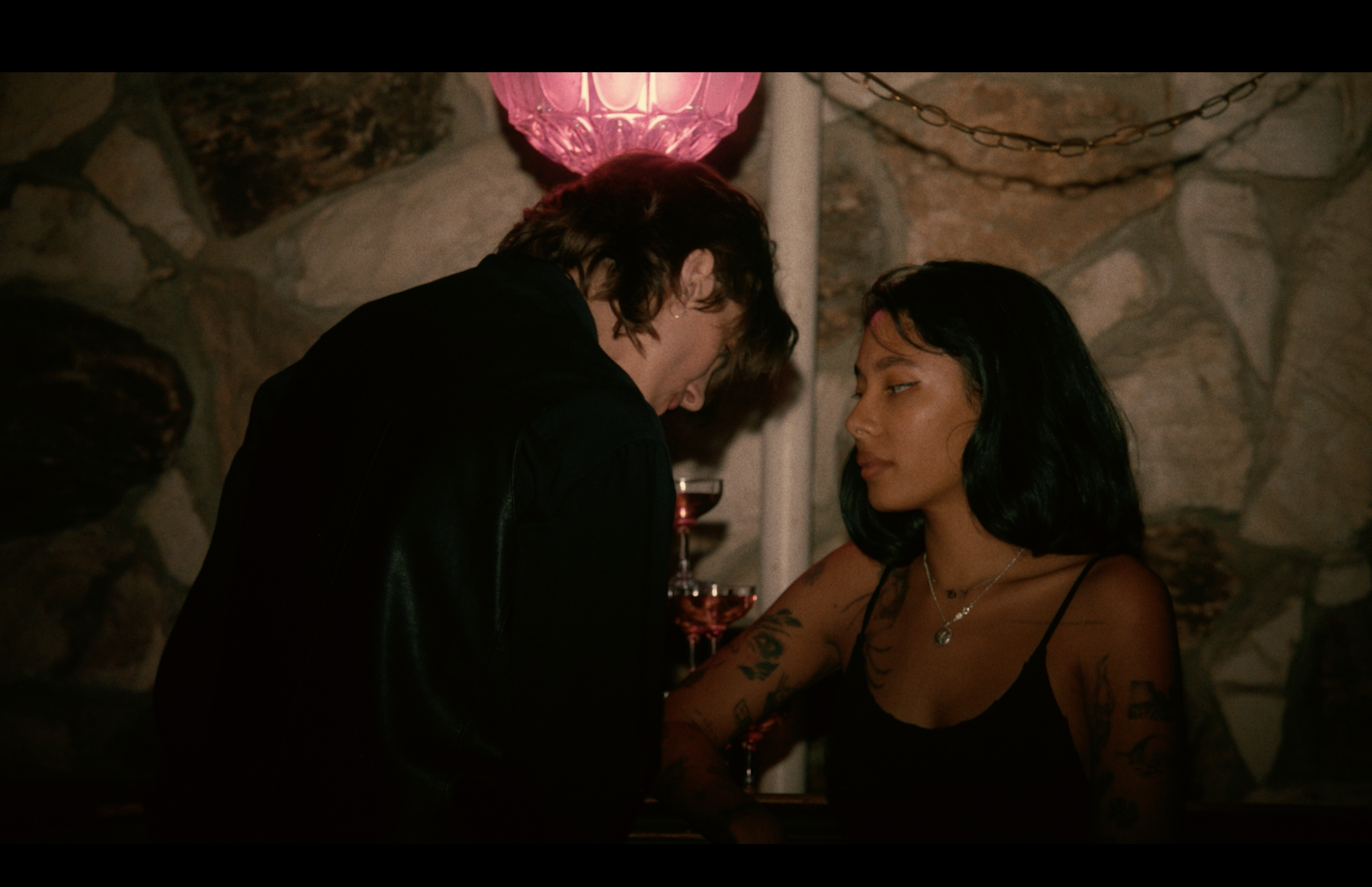 Two women are in a dimly lit room, leaning close and sharing an intimate moment. One woman has short dark hair and tattoos on her arms, wearing a black top, and the other has medium-length dark hair and is wearing a black spaghetti strap top. They ar