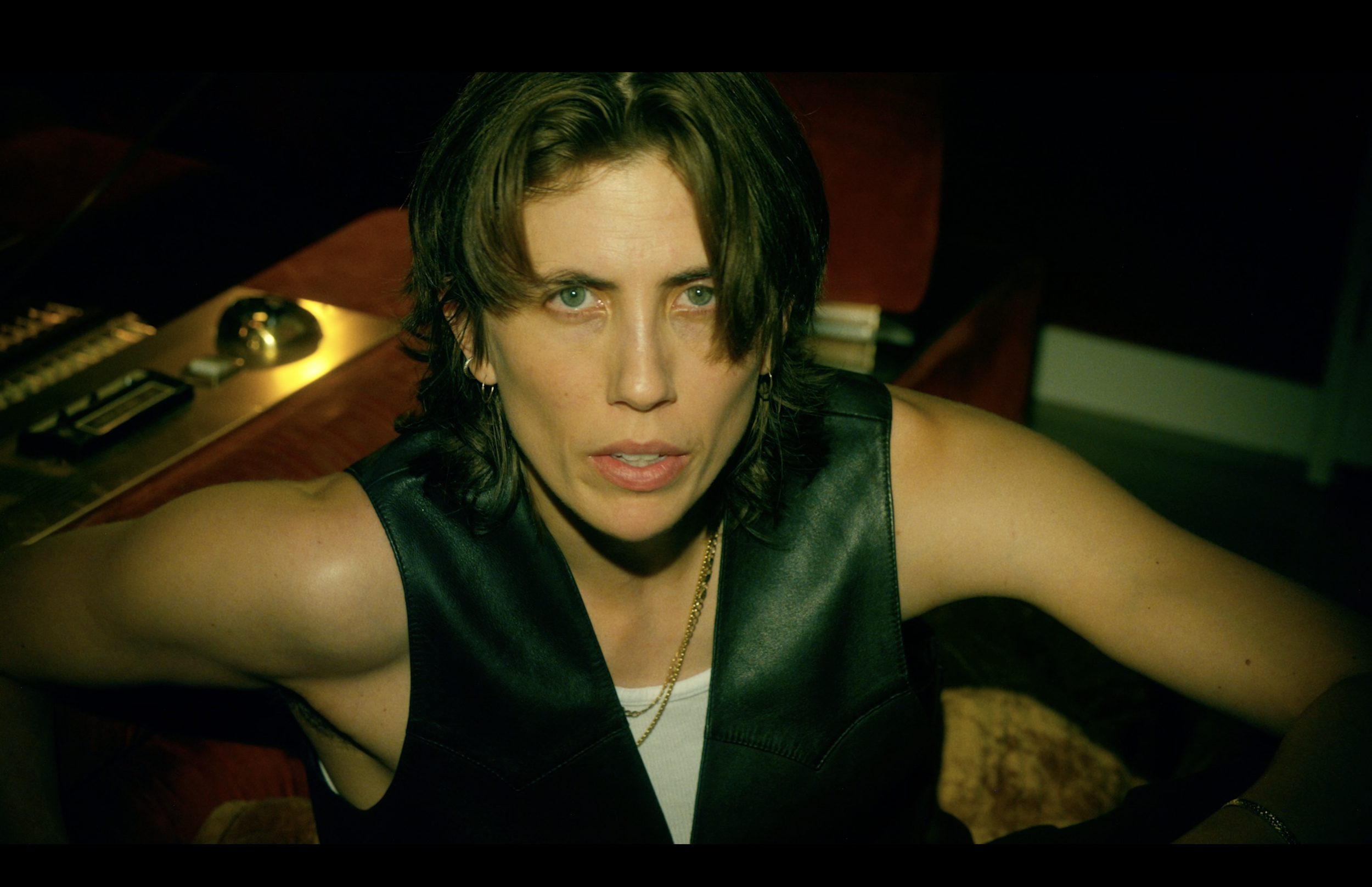 A woman with short dark hair and blue eyes wearing a black leather vest, sitting at a table with a serious expression.